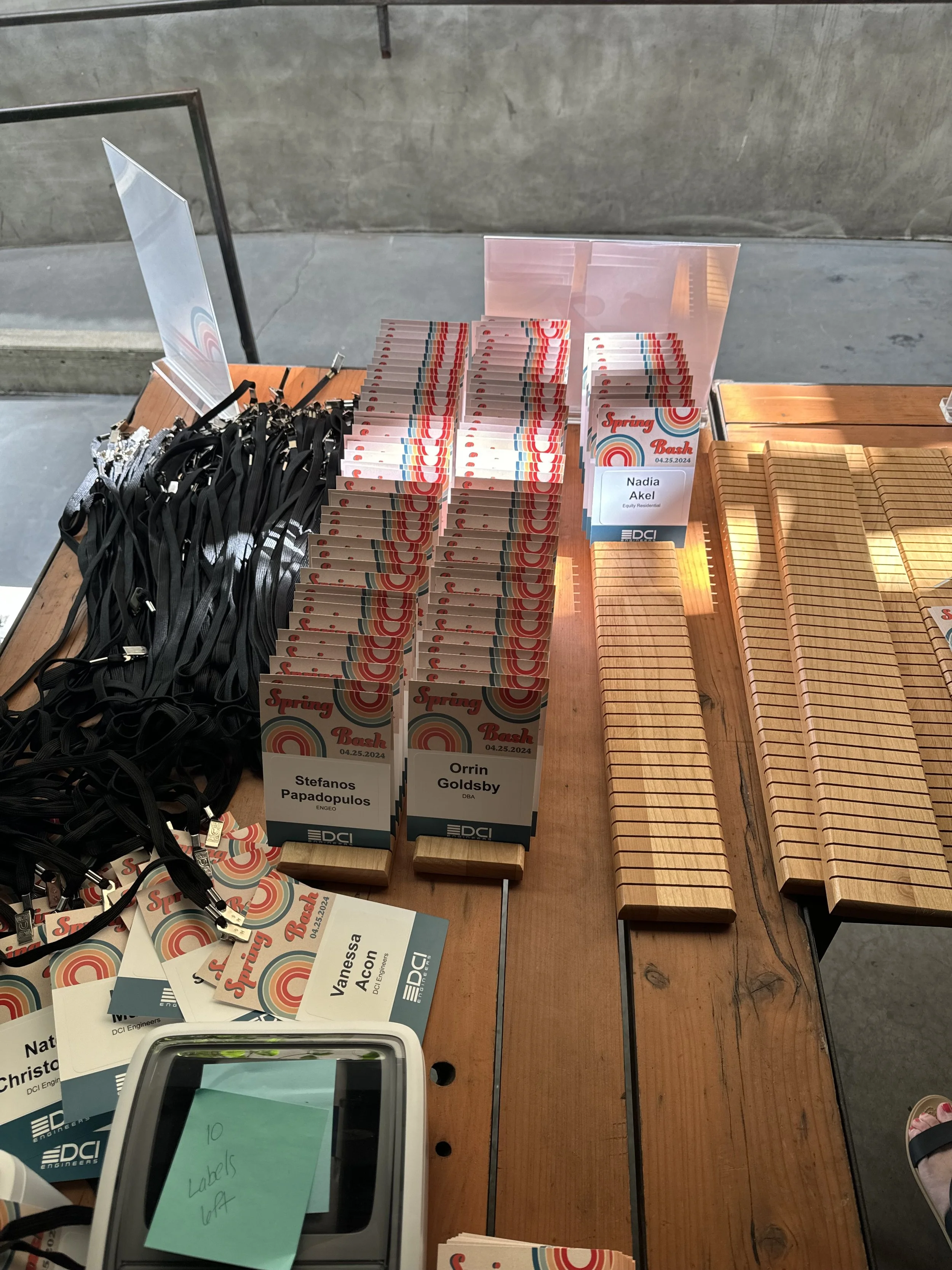 Conference registration table with name tags, lanyards, and wooden name plate holders at an event. Some name tags are visible, including one for 'Nadia Akel' and another for 'Orrin Goldsby'. The table also has a note that says '10 Labels U4'.