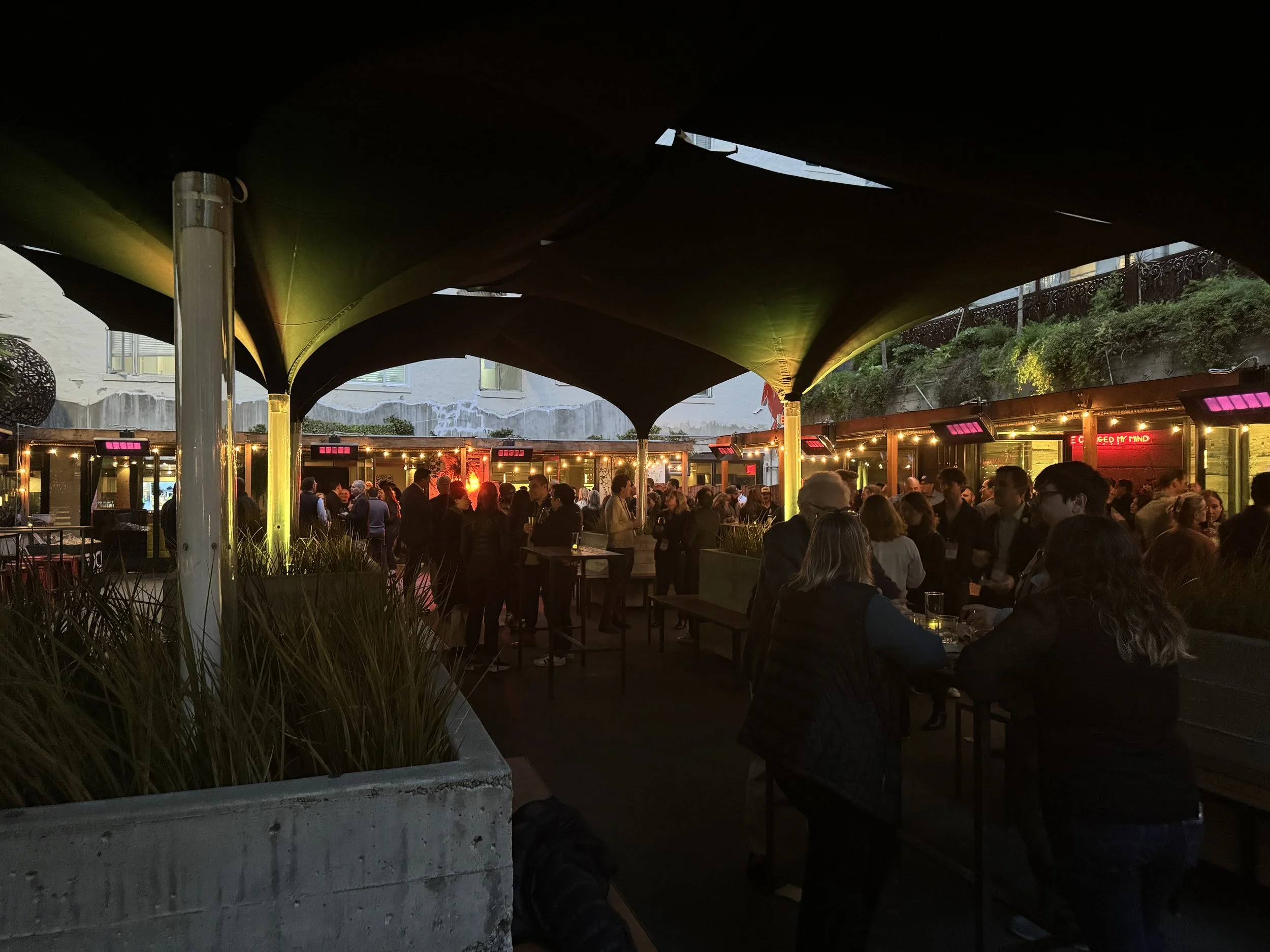 Outdoor bar and patio with a crowd of people socializing under a large canopy at night, with warm lighting and neon signs.