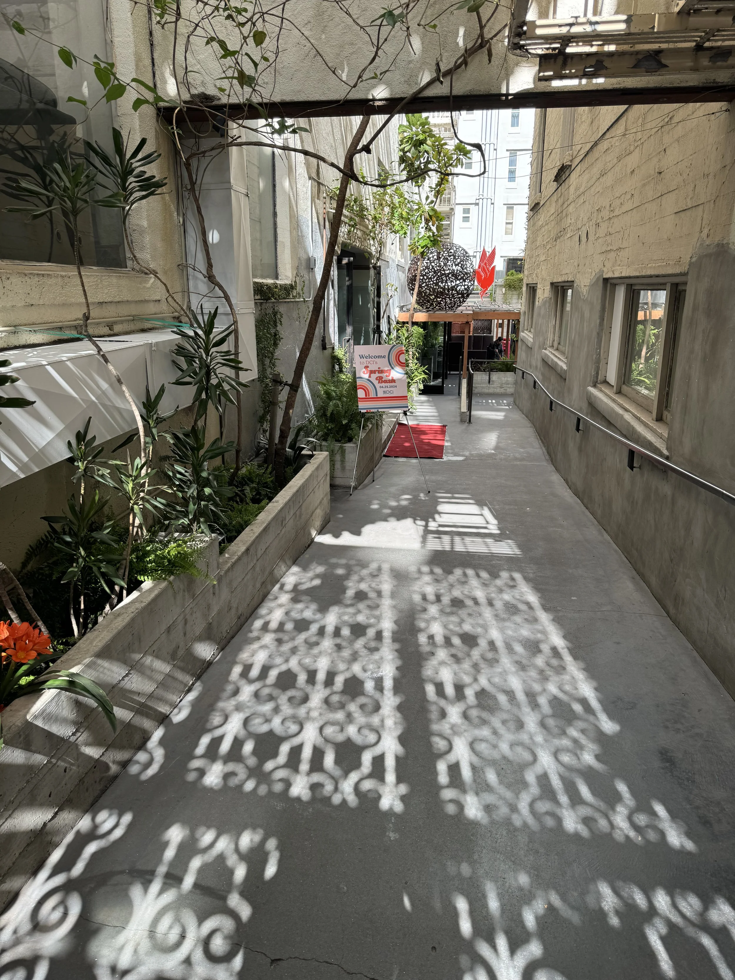 Urban alleyway with plants in concrete planters on the left and a textured shadow cast black iron fence on the ground. A sign, a red welcome mat, and decorative sculptures are visible at the end of the alley.
