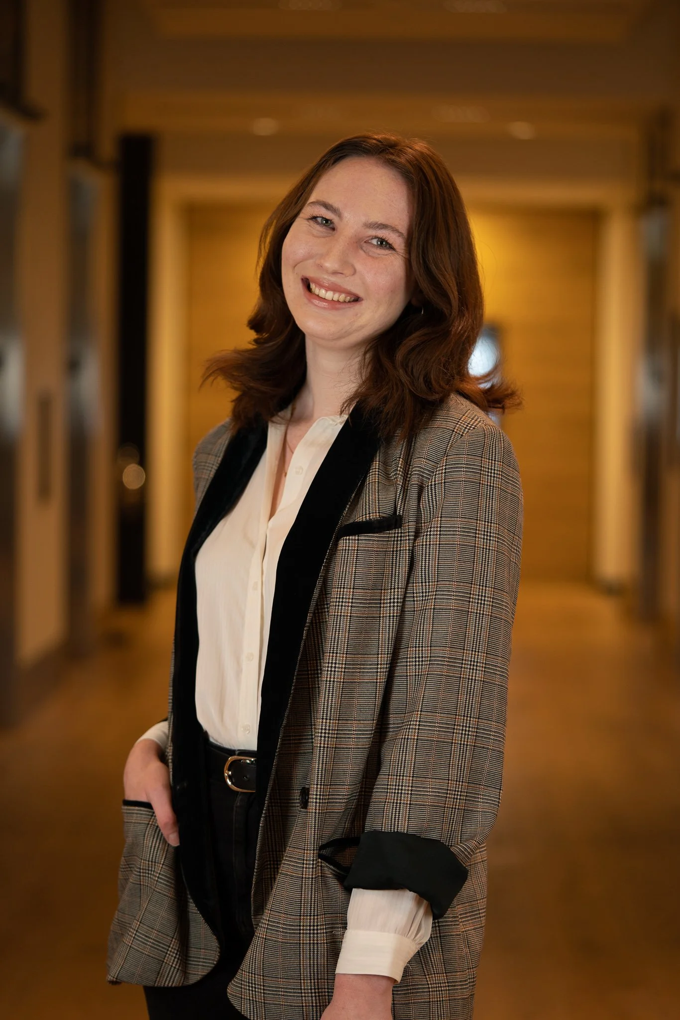 A young woman with shoulder-length brown hair, smiling, wearing a plaid blazer over a white blouse, standing in a hallway with warm lighting.