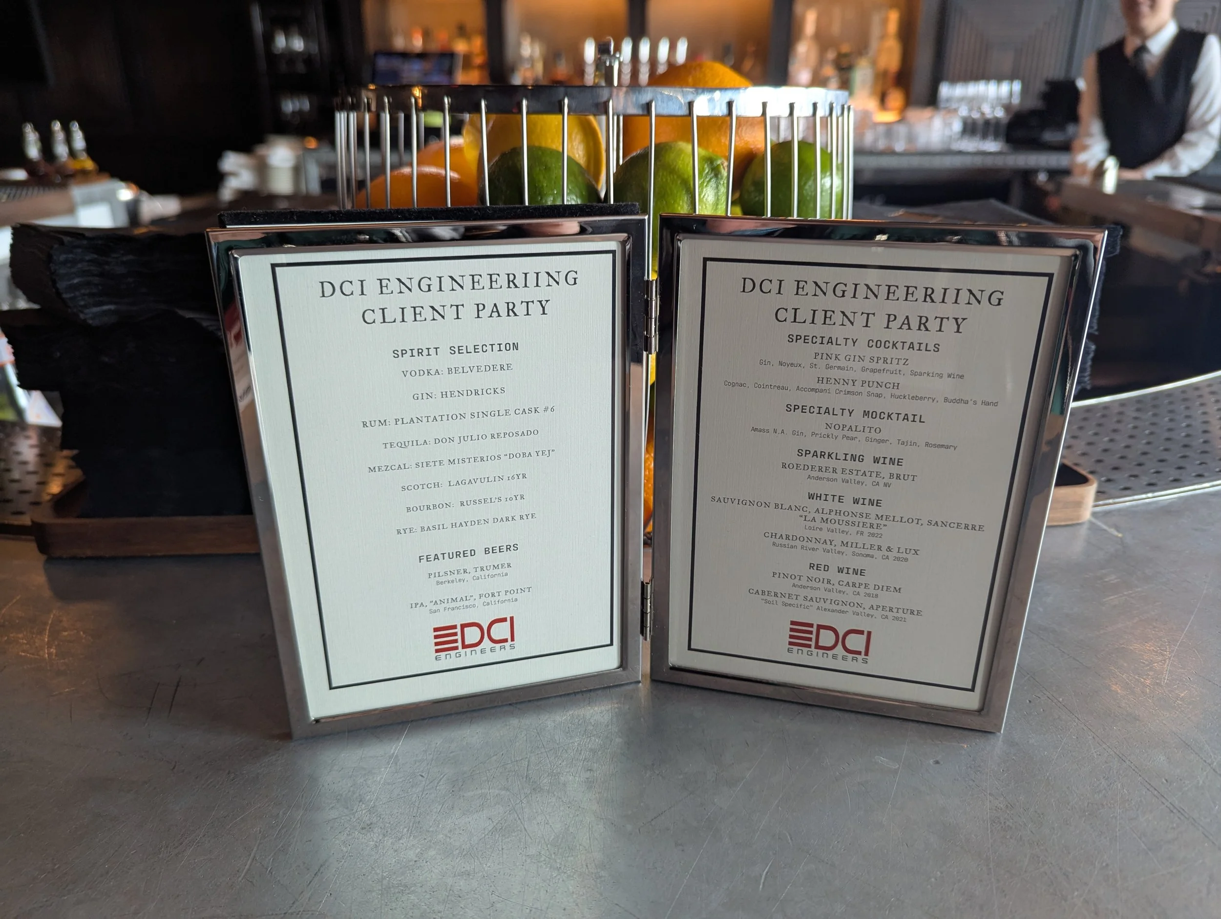 Two drink menu cards on a table at a bar for DCI Engineering Client Party featuring spirit selection and specialty cocktails, with a metal fruit basket in the background.