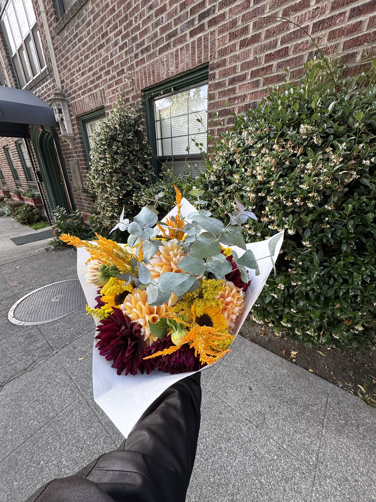 A person holding a colorful bouquet of flowers on a city sidewalk with brick buildings and greenery in the background.