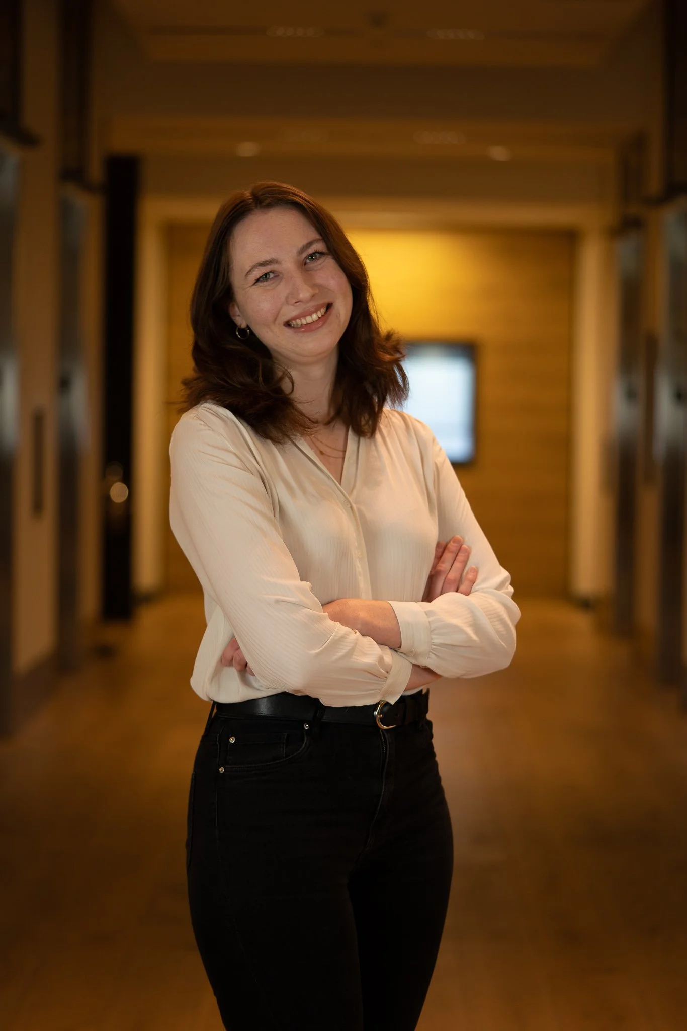 A woman with shoulder-length brown hair, wearing a cream-colored blouse and black jeans, smiling with arms crossed, standing in a warmly lit wooden hallway.