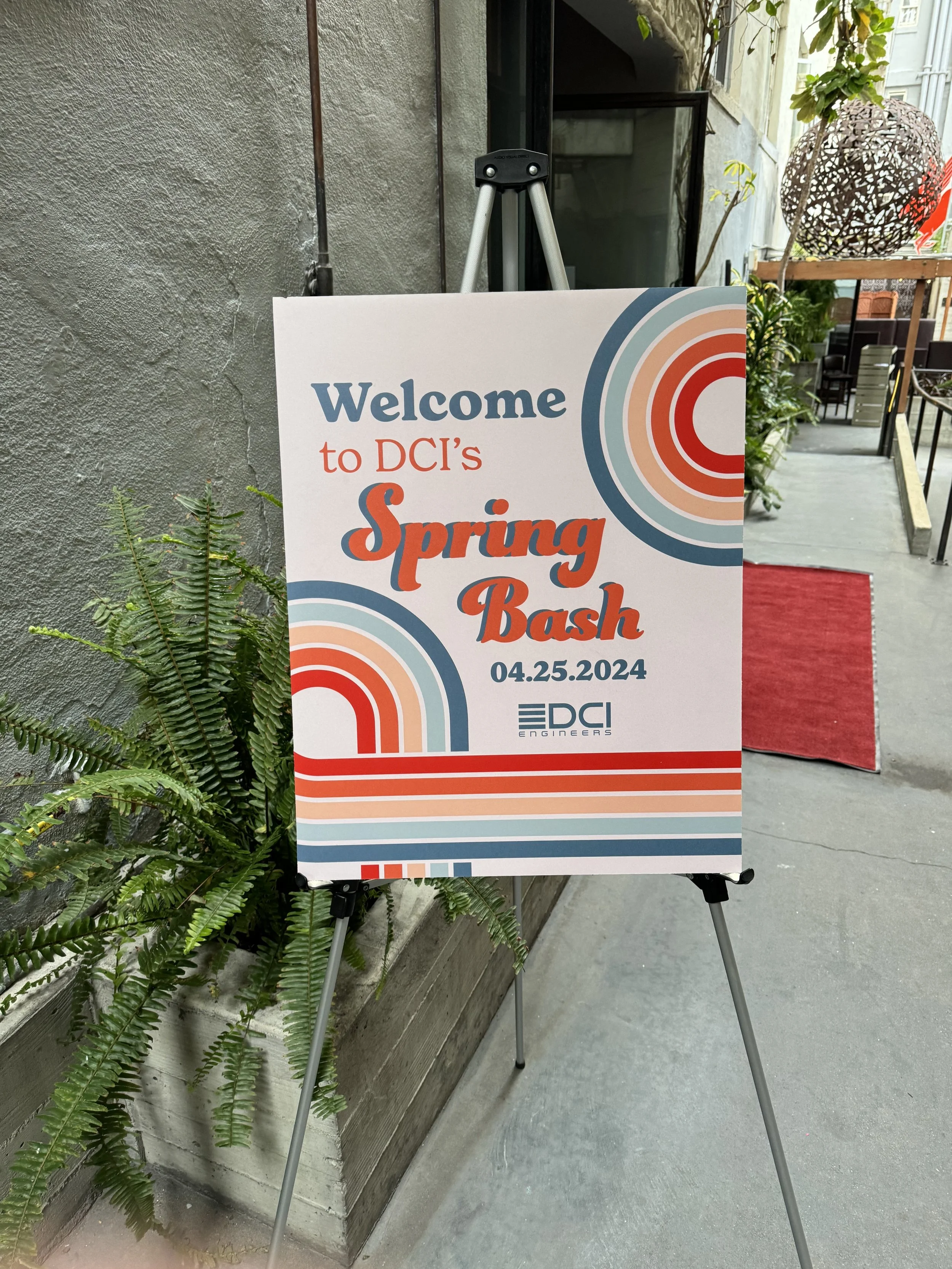 Signboard welcoming attendees to DCI's Spring Bash on April 25, 2024, with colorful retro-style design, placed outside on a sidewalk next to some plants and a building.