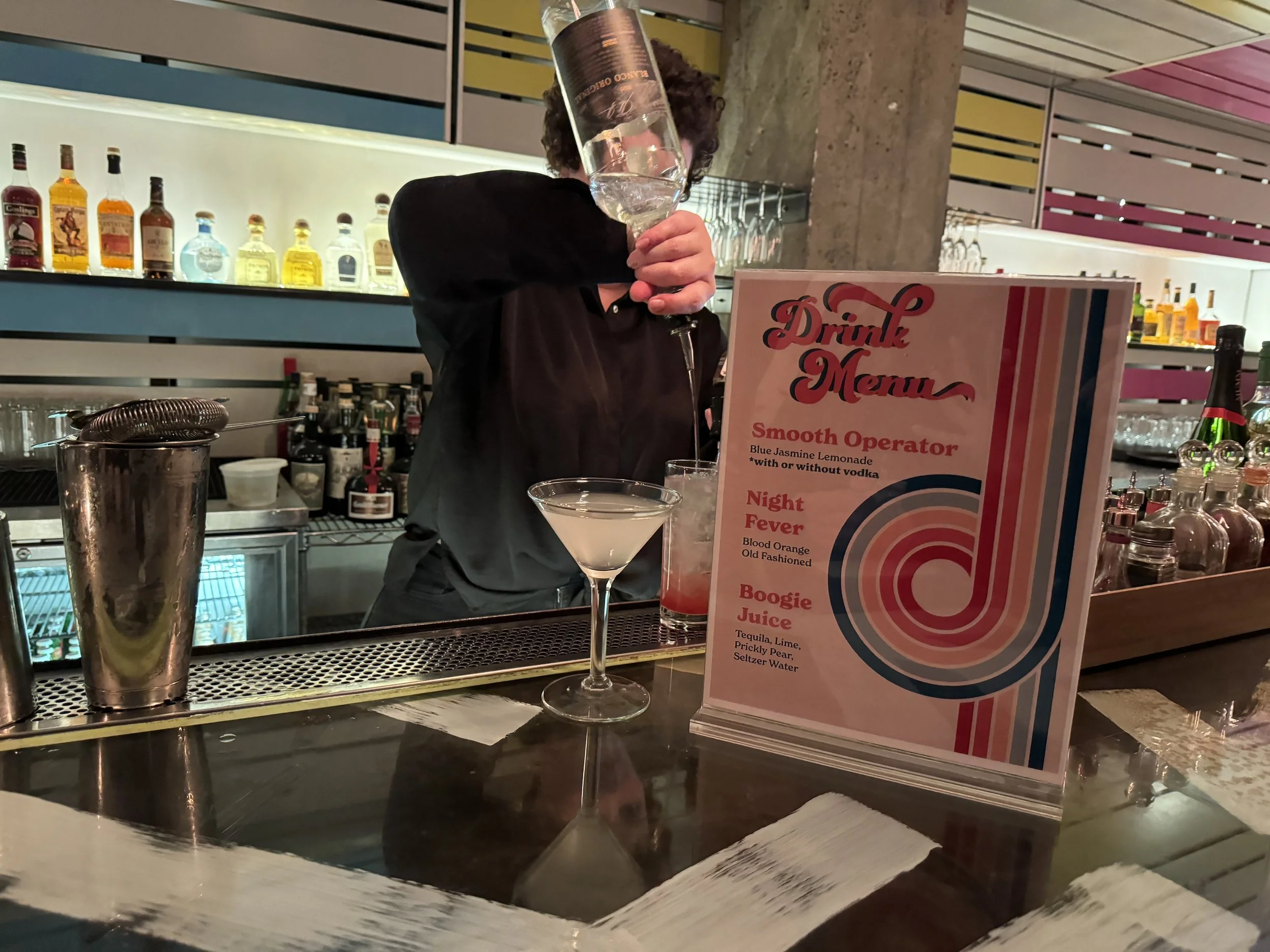 Bartender pouring a cocktail into a glass at a bar with a drink menu in the foreground.