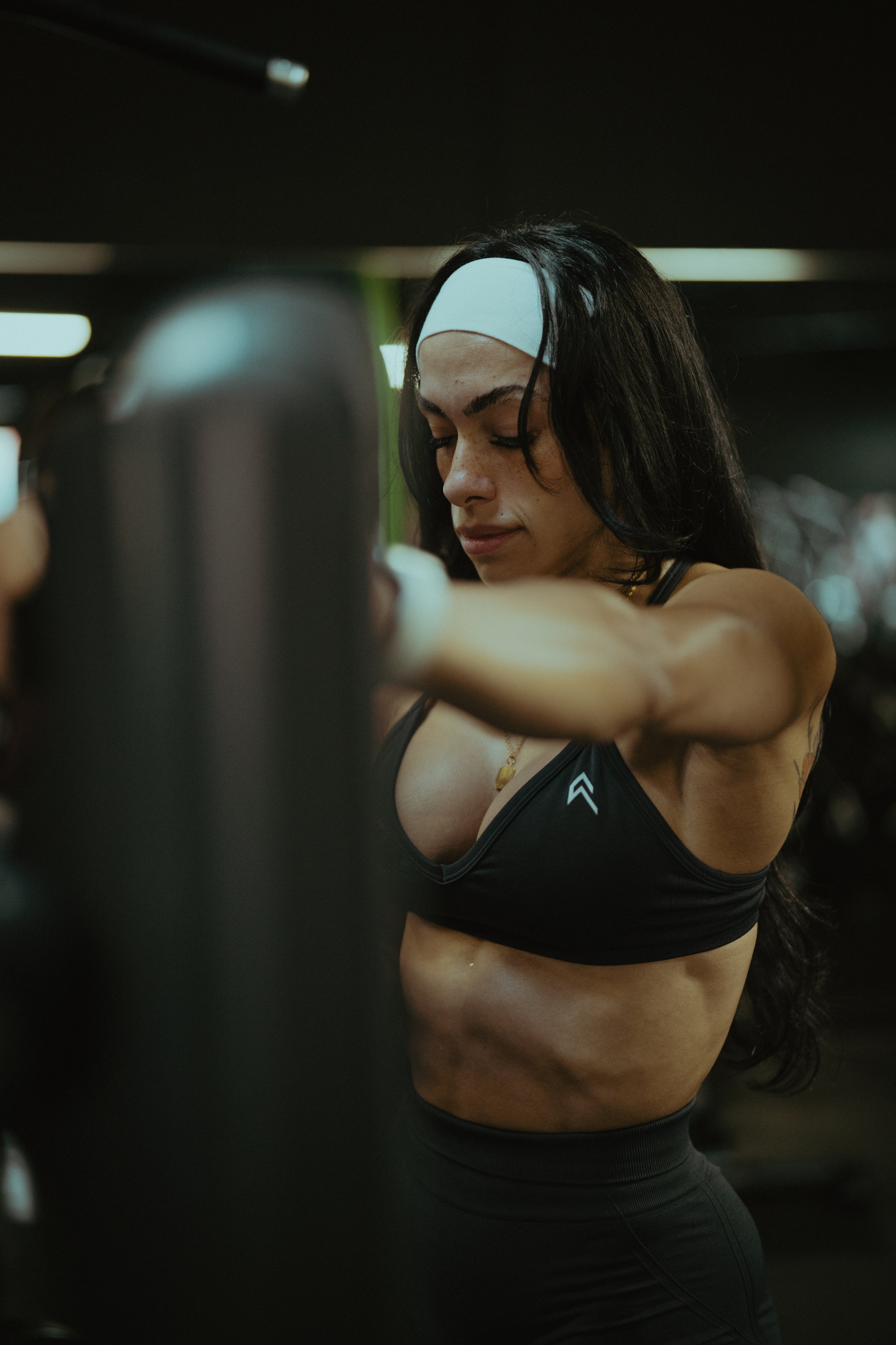 A woman with long dark hair wearing a white headband, black sports bra, and black workout leggings, stretching her arms at the gym.