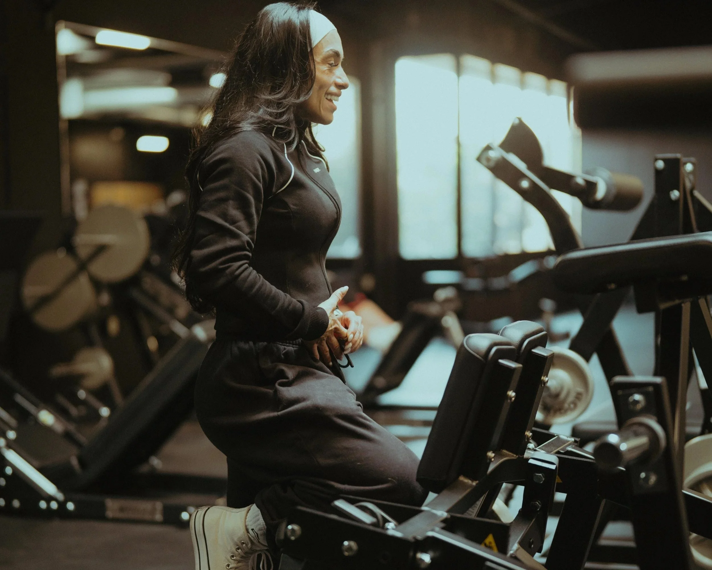 A woman in a black working out gear using a leg press machine in a gym, smiling, with sunlight coming through large windows in the background.
