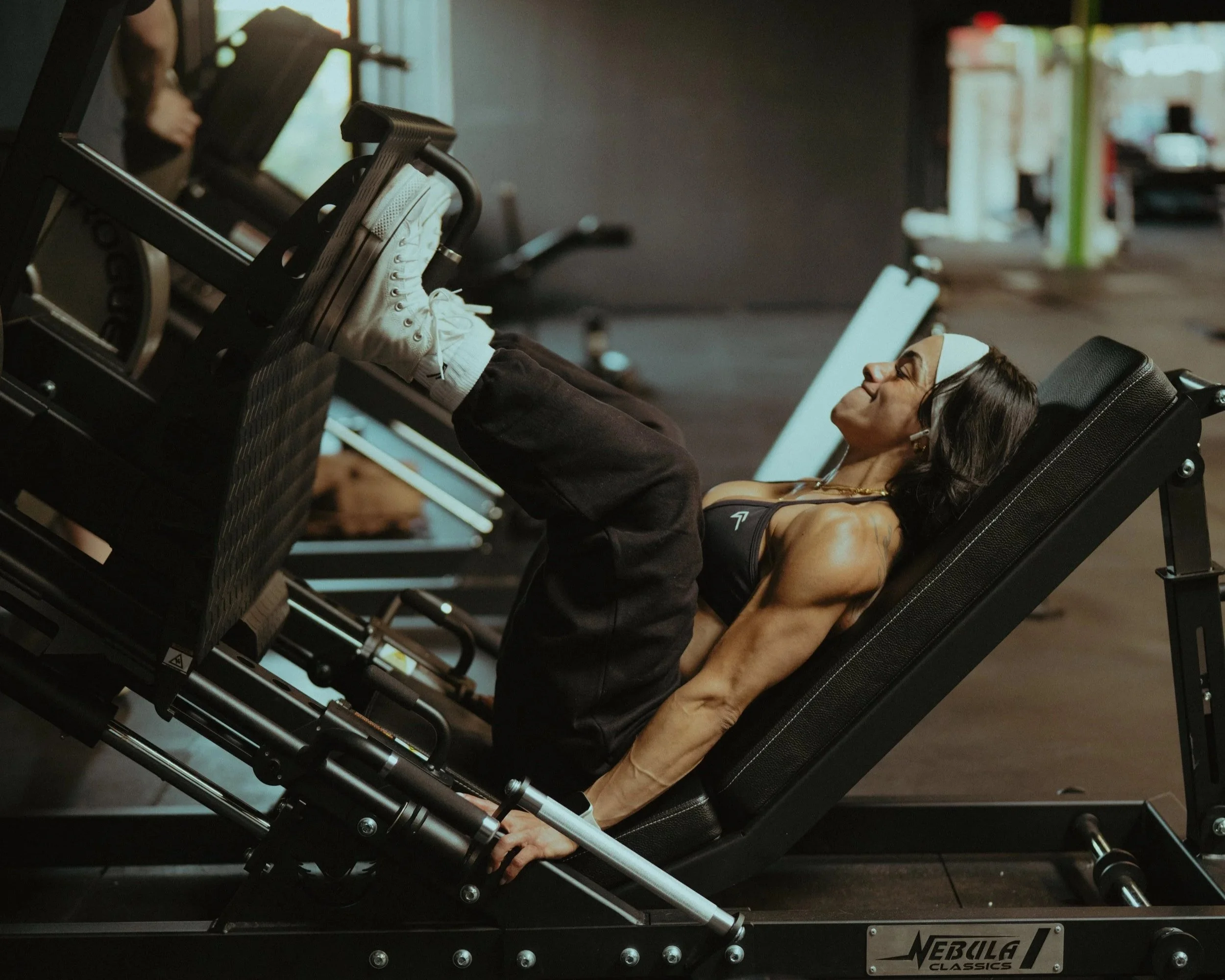 A woman with toned muscles lying on a fitness machine in a gym, wearing a sports bra and headband.