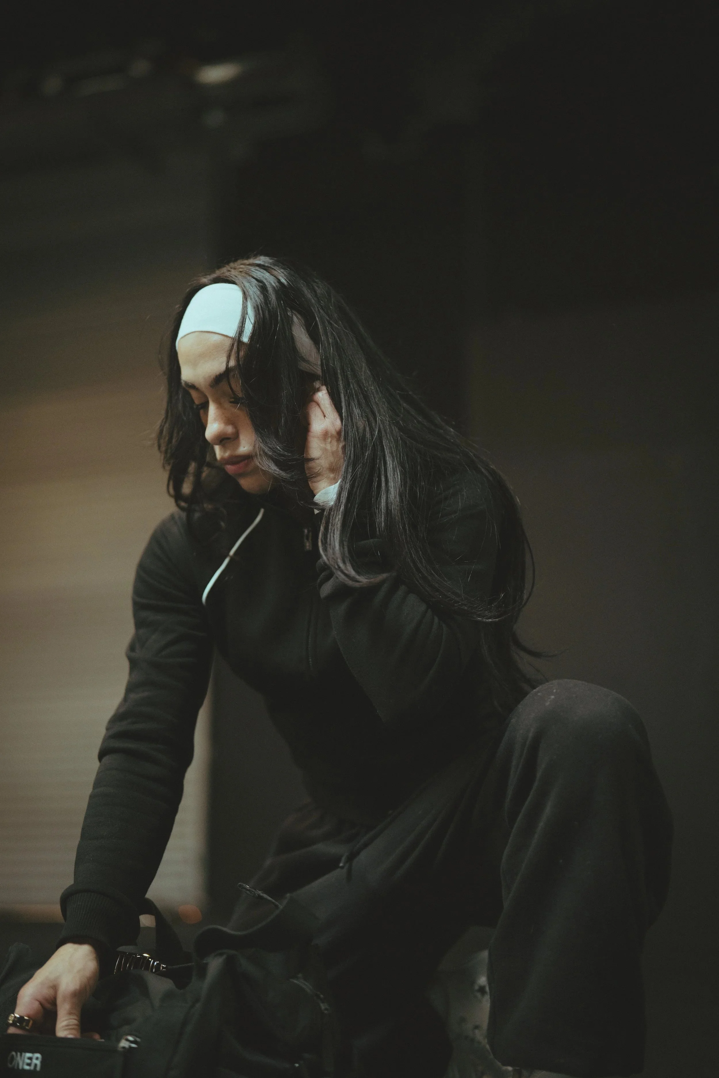 Woman with long black hair, wearing a black jacket, touching her ear, in a dark indoor setting.