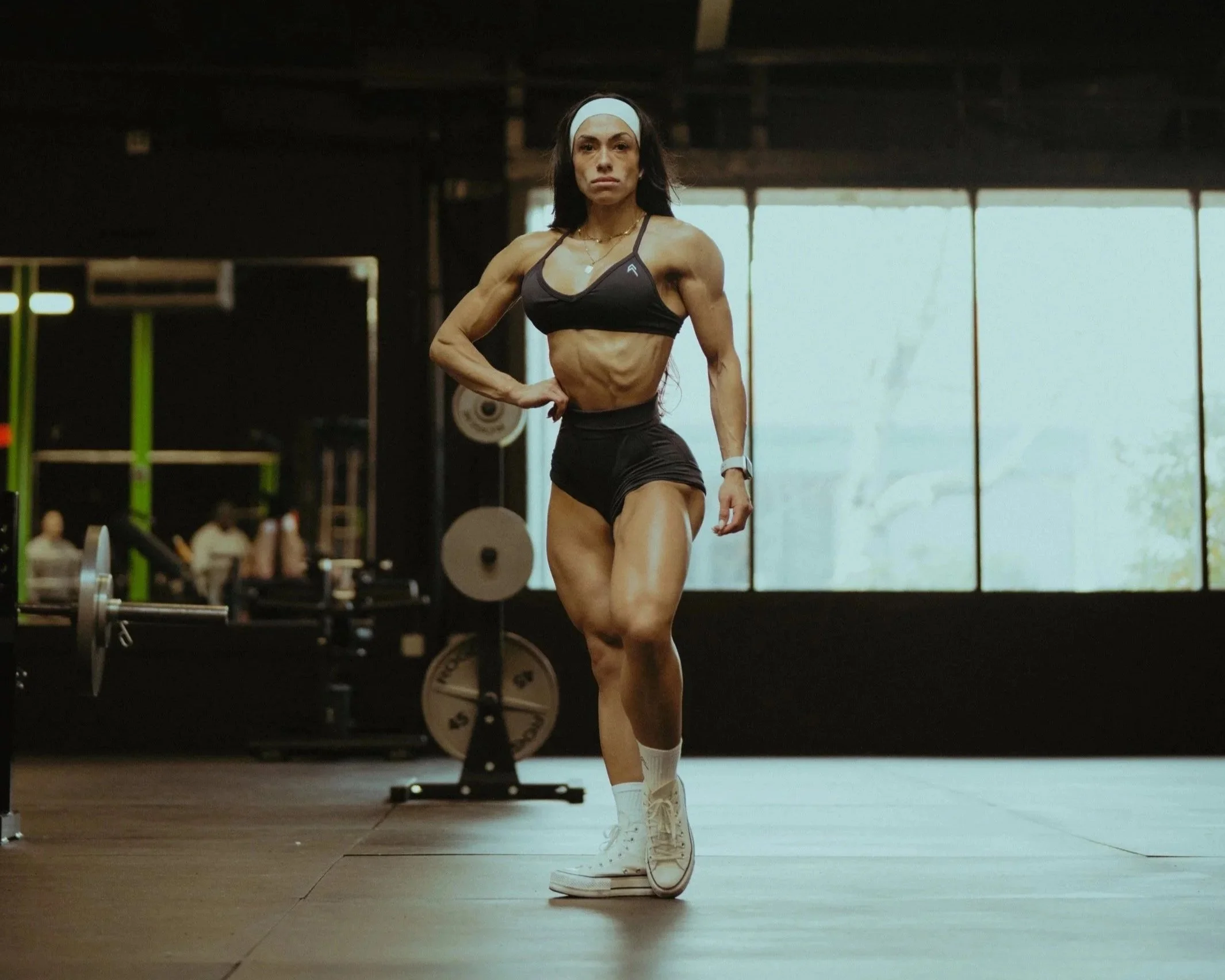 A fit woman in gym attire stands confidently in a gym, with one hand on her hip, surrounded by weightlifting equipment and large windows showing daylight outside.