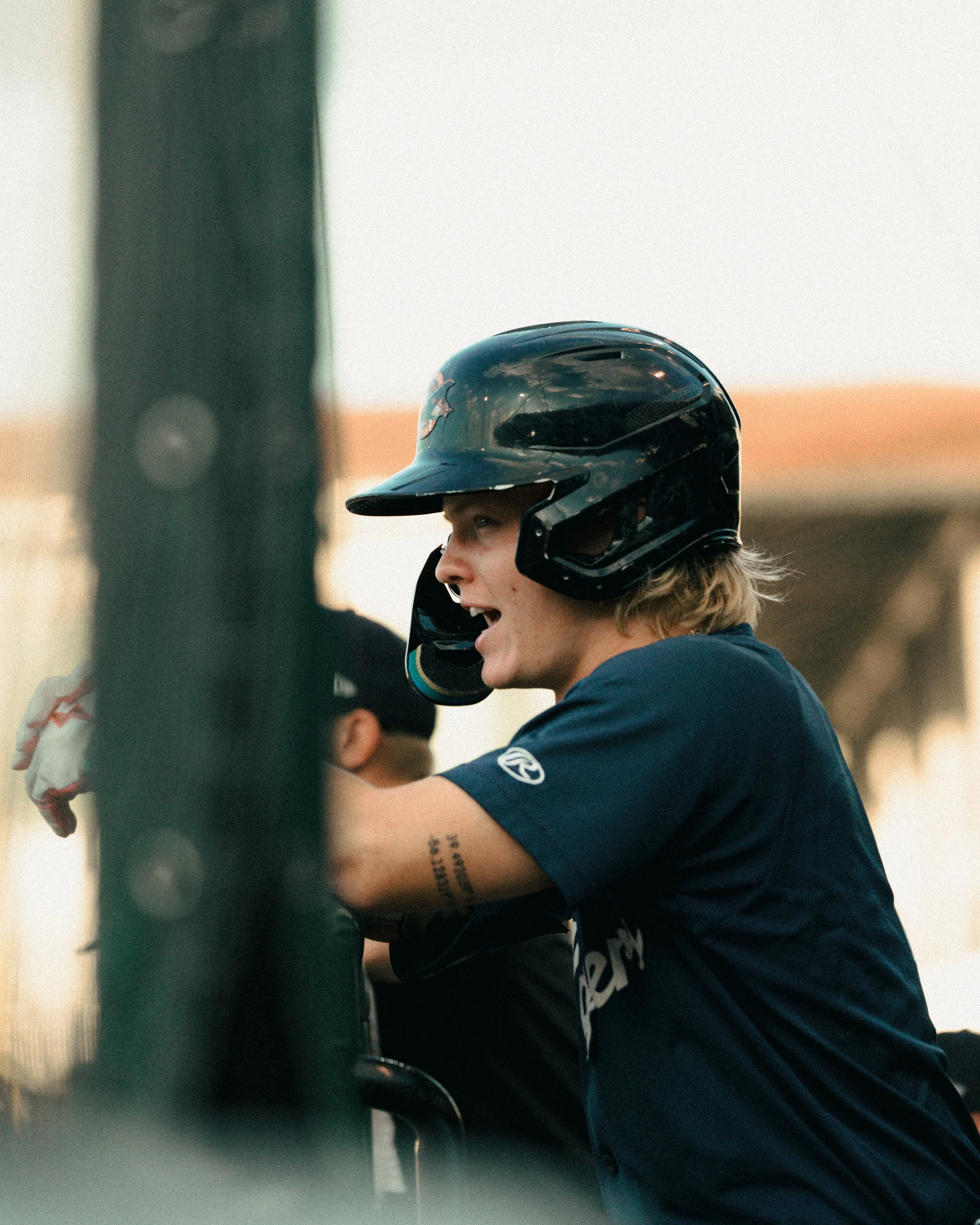 A young man with blond hair wearing a black helmet and a blue race jersey, leaning on a motorcycle during a race or practice session at an outdoor track.