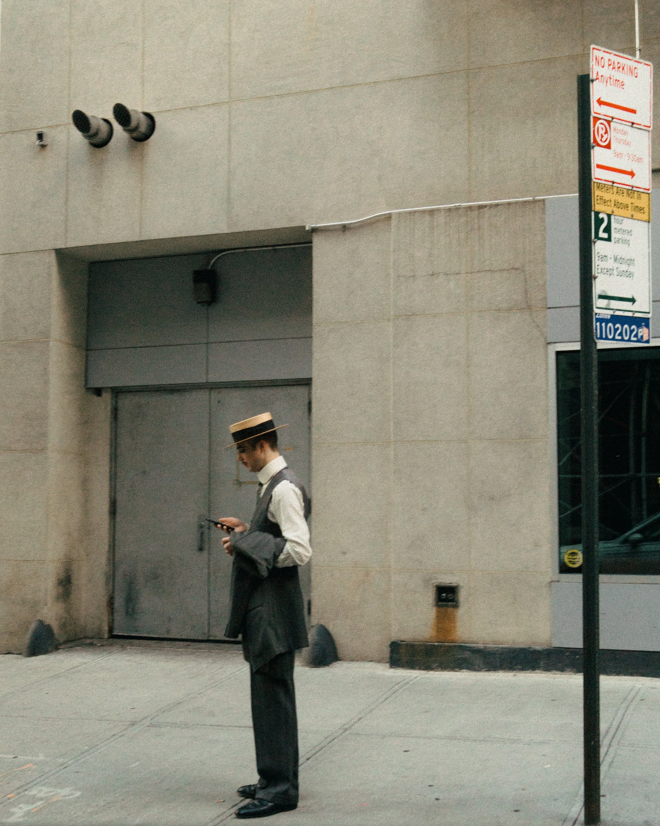 A man dressed in vintage clothing, wearing a straw boater hat, standing on a city sidewalk and looking at his phone.