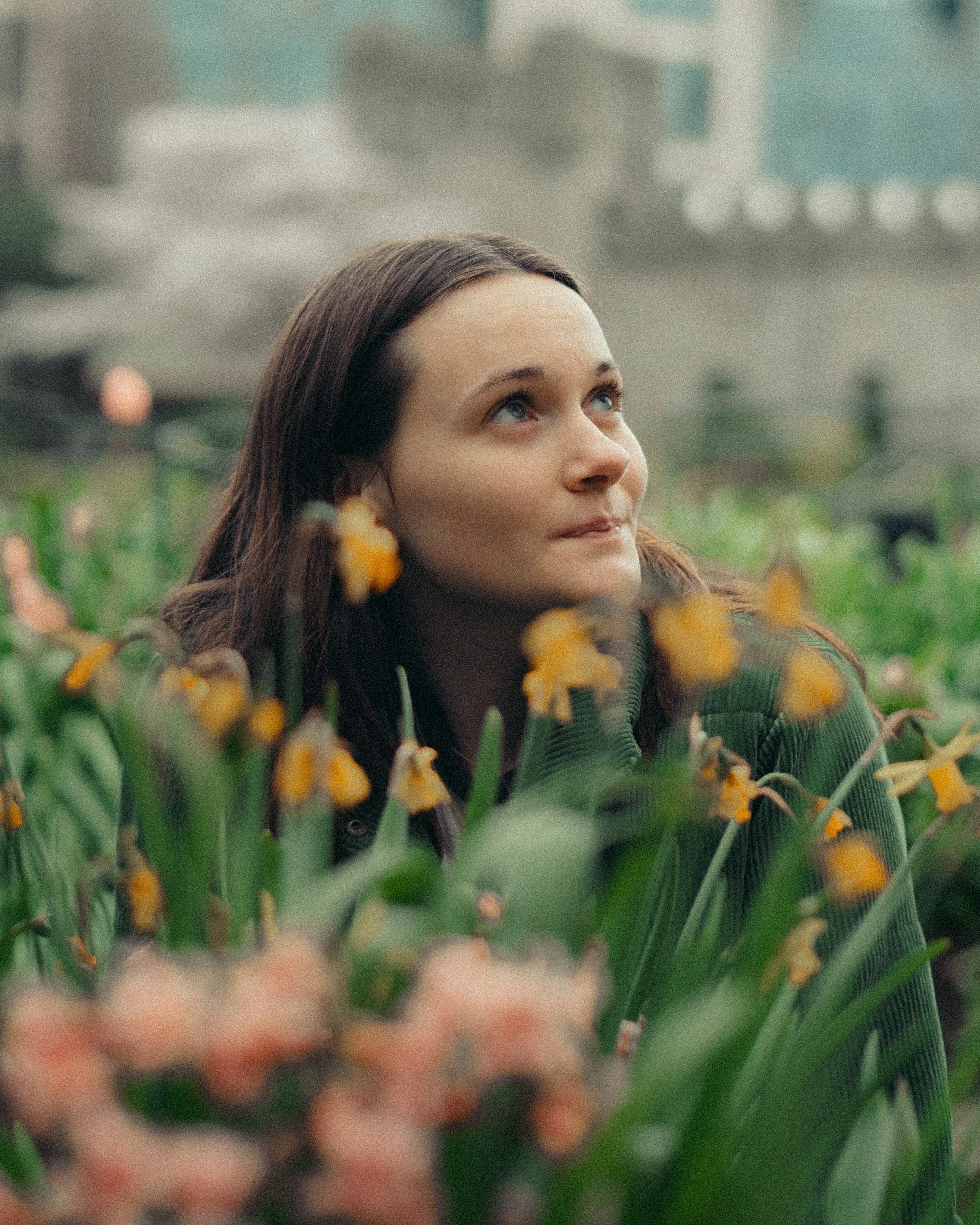 A woman with long brown hair and blue eyes looking up among yellow flowers in a garden with city buildings in the background.