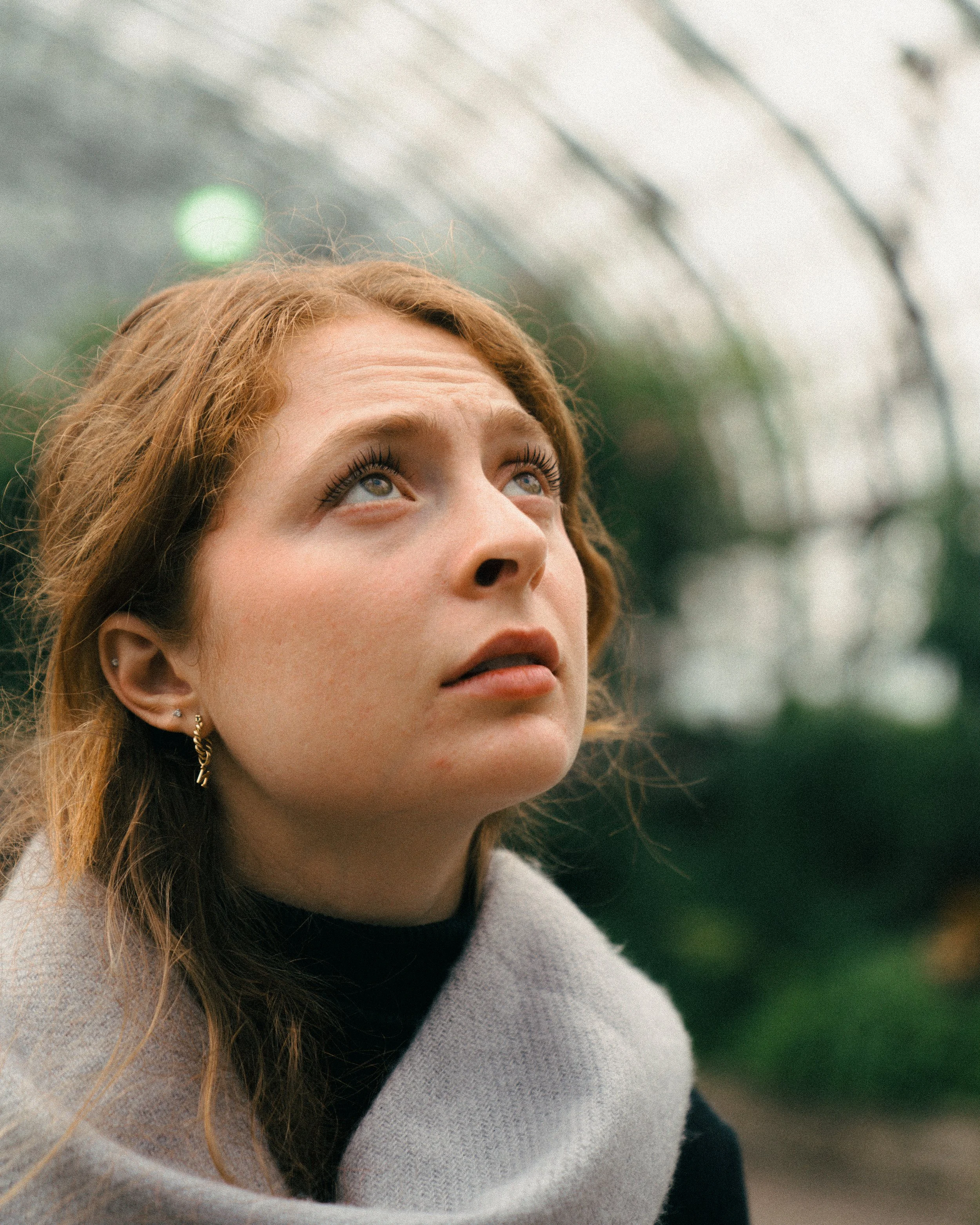Close-up of a woman with red hair looking up, inside a greenhouse or conservatory with a glass ceiling
