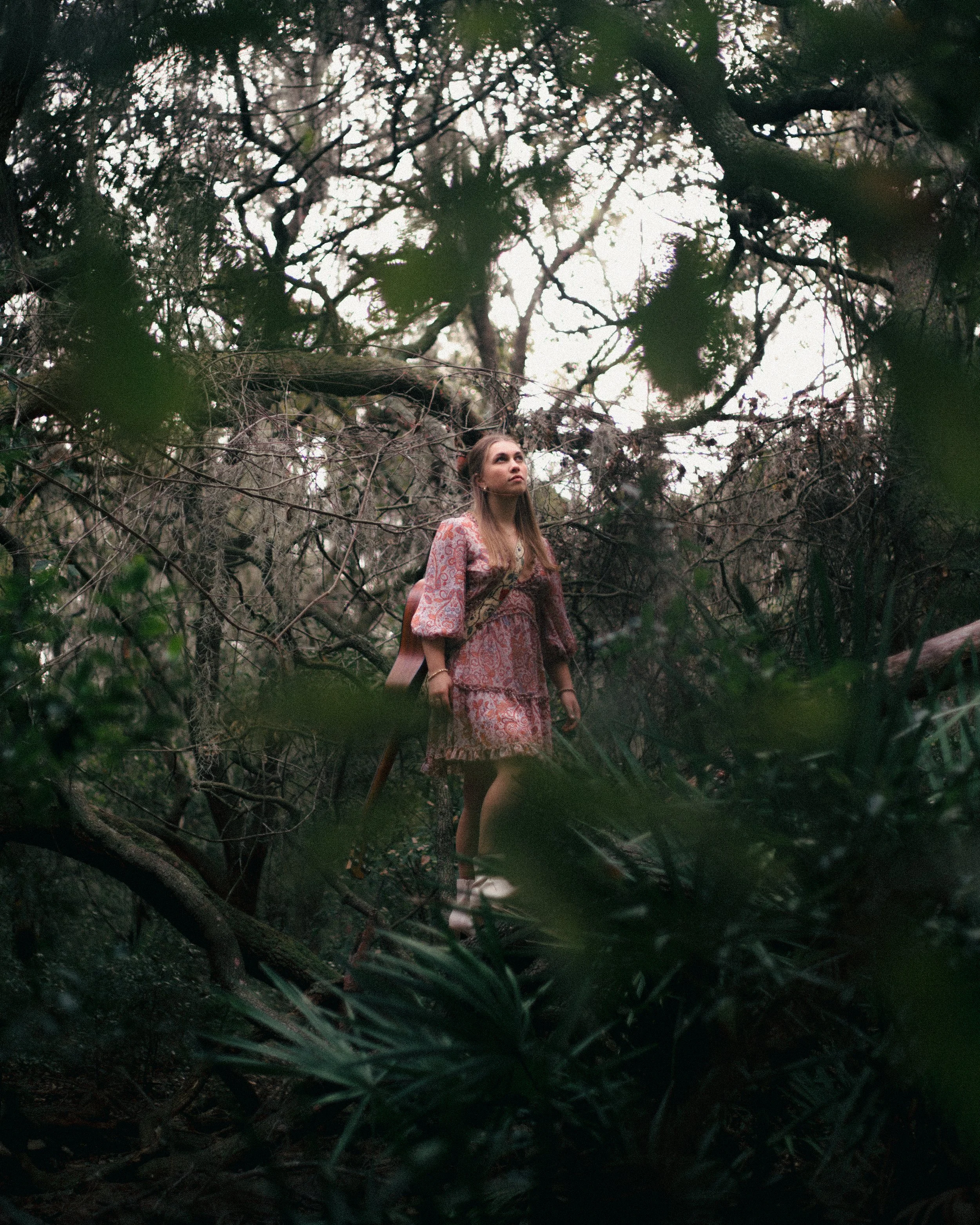 Young woman with long hair standing in a dense, dark forest, looking upwards, dressed in a patterned pink and brown dress.
