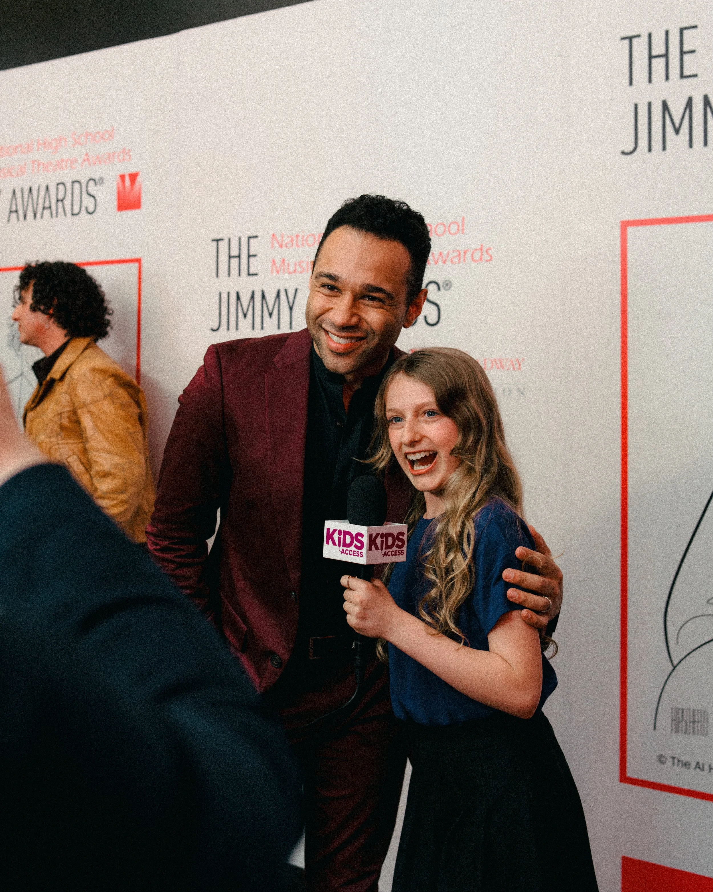 A man in a maroon suit and a girl with long blonde hair holding a Kids' Access microphone, smiling and posing for a photo at an award event with a backdrop that has text about 'The Jimmy' awards.