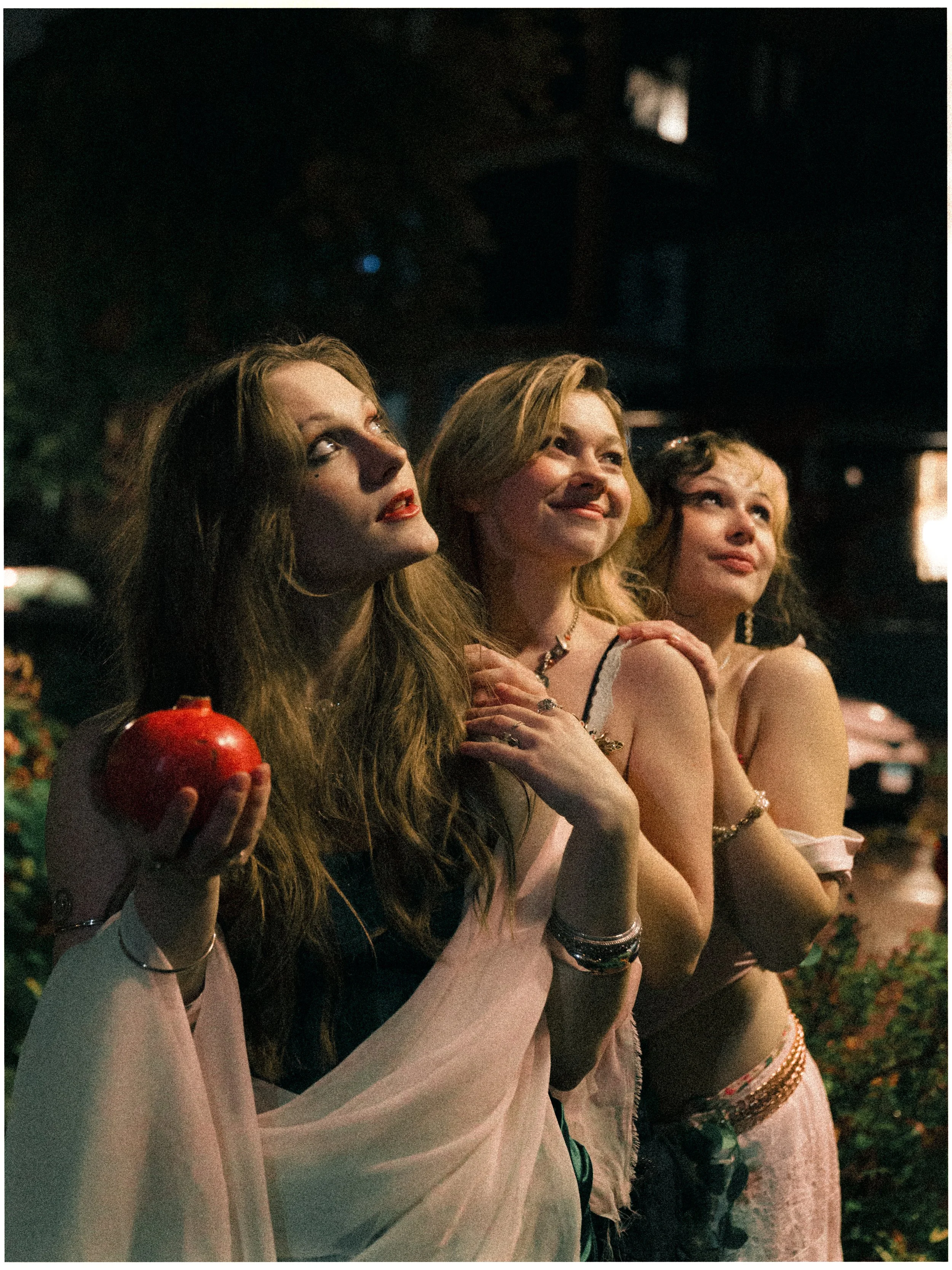 Three women standing outdoors at night, dressed in party or costume attire. The woman on the left is holding a red apple, has long reddish-brown hair, and appears contemplative. The woman in the middle has long blonde hair and is smiling softly. The 