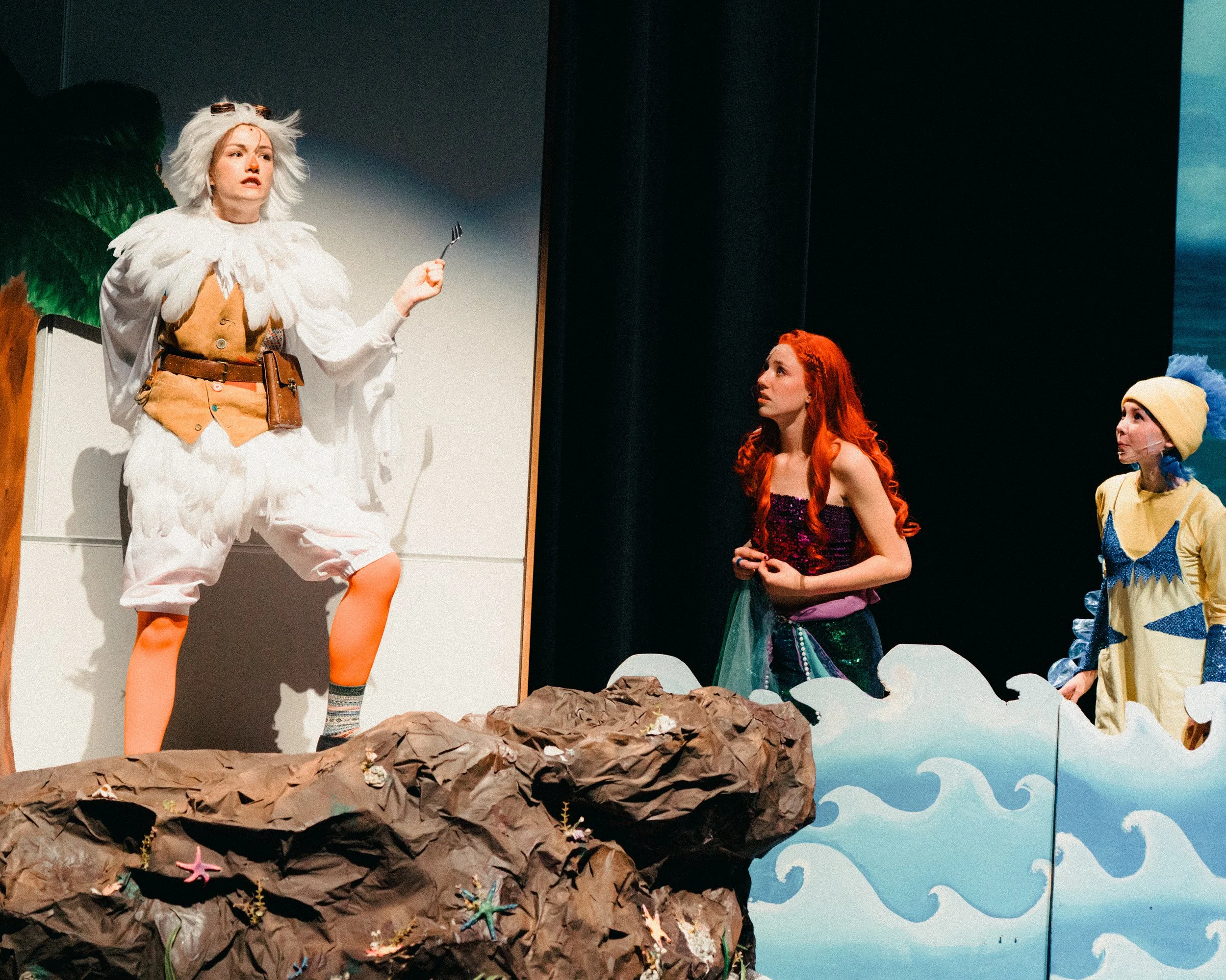 Three actors performing in a theatrical play on stage, with one dressed as an outdoor explorer, and two dressed as animated characters, one with red hair and the other with a yellow headpiece, against a backdrop of painted scenery.