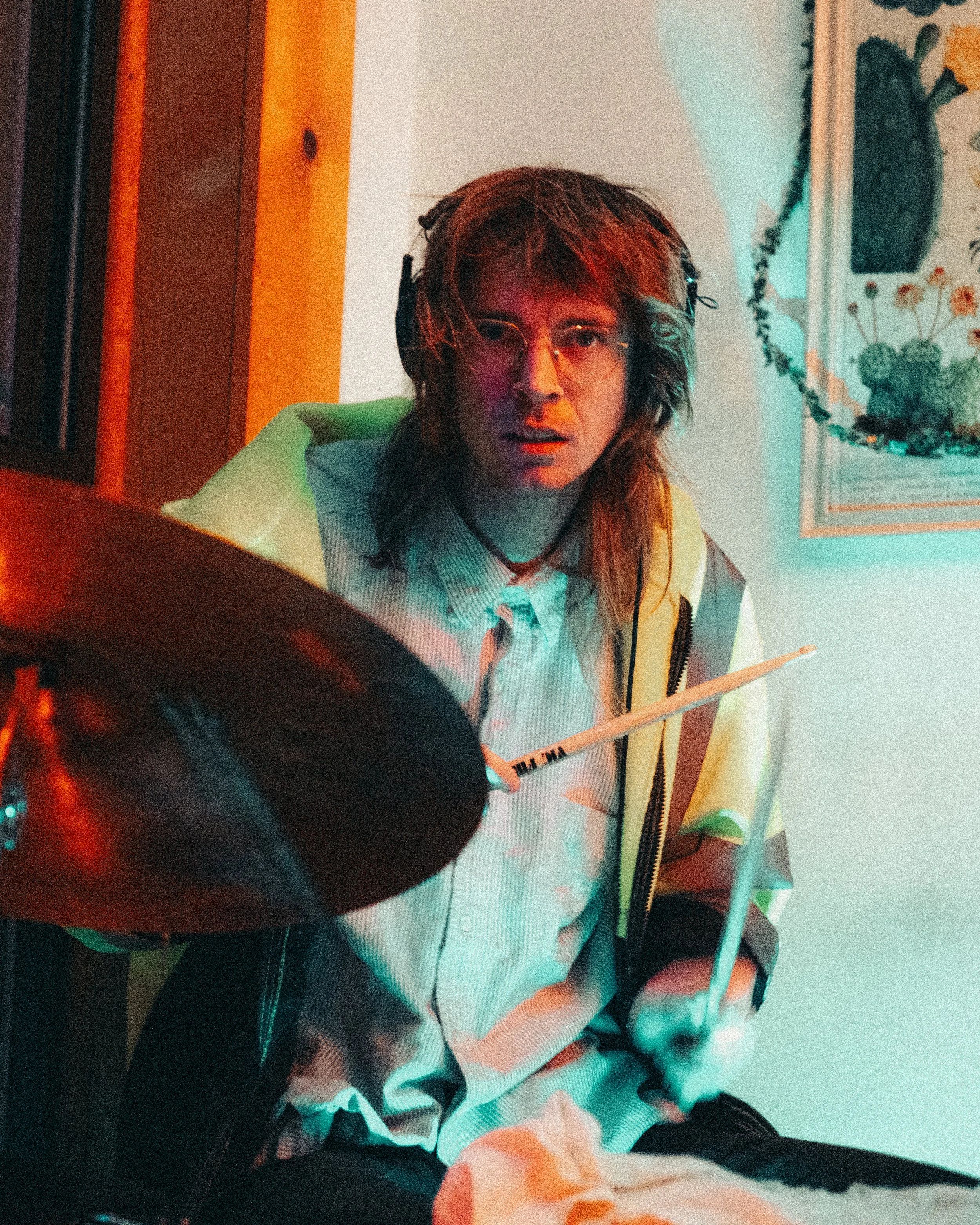 A young person with long hair and glasses, wearing a striped shirt and headphones, sitting in front of a drum set, holding drumsticks.