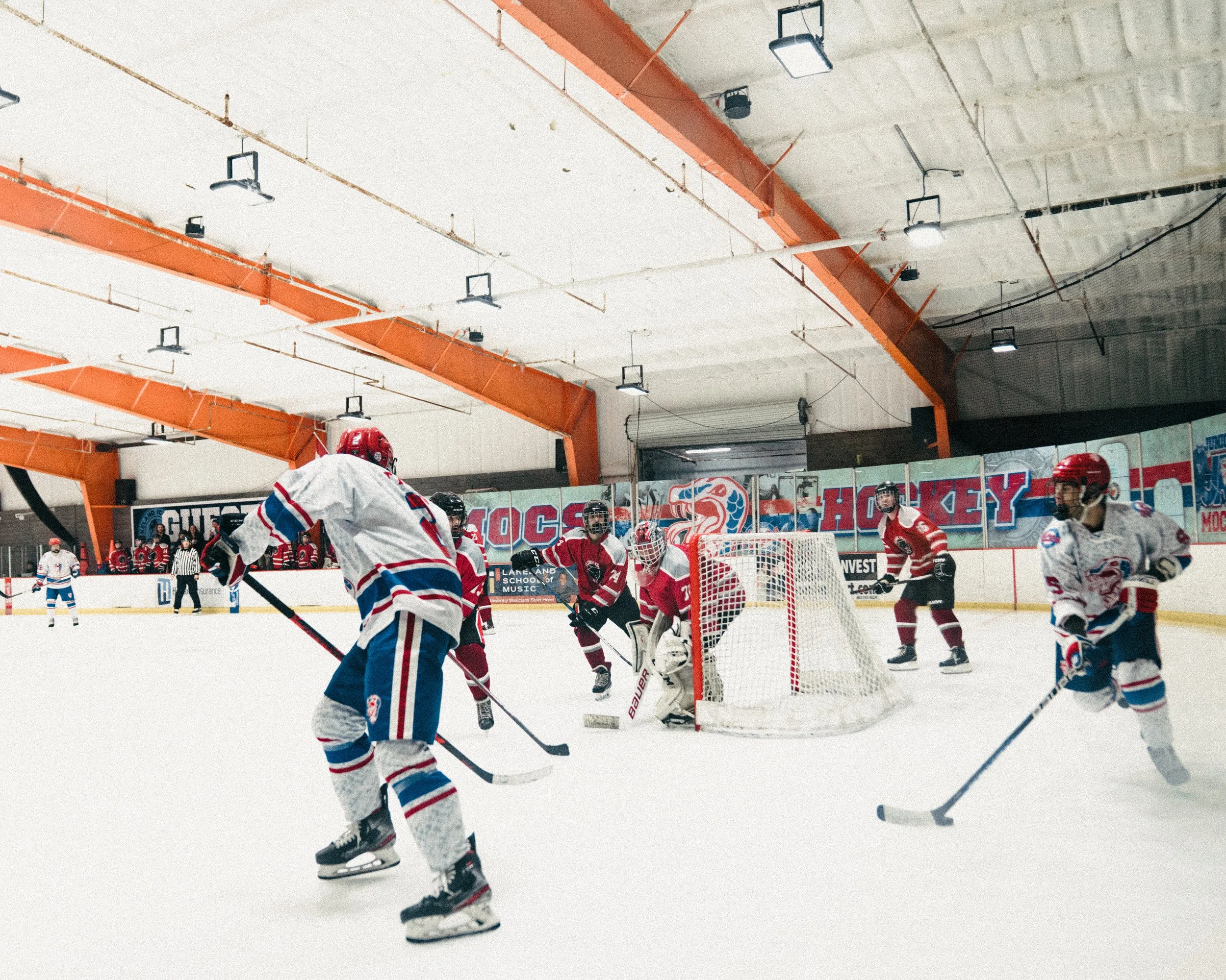 Ice hockey game in progress with players in red and white jerseys near the goal, some players skating, and a referee in the background, inside an indoor ice rink with orange beams overhead.