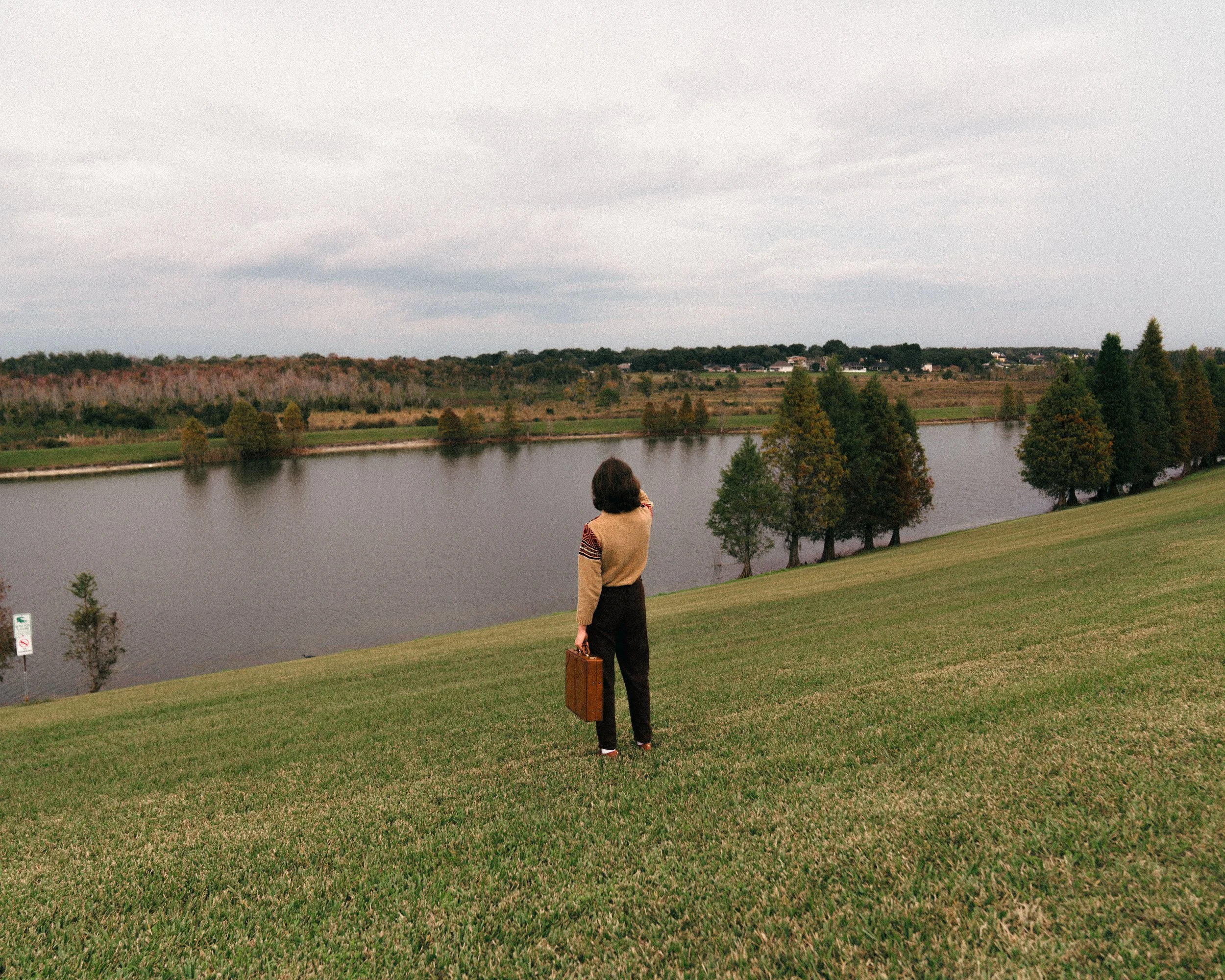 A woman in a beige and brown sweater standing on a grassy hill near a body of water, holding a brown suitcase, with trees and an overcast sky in the background.