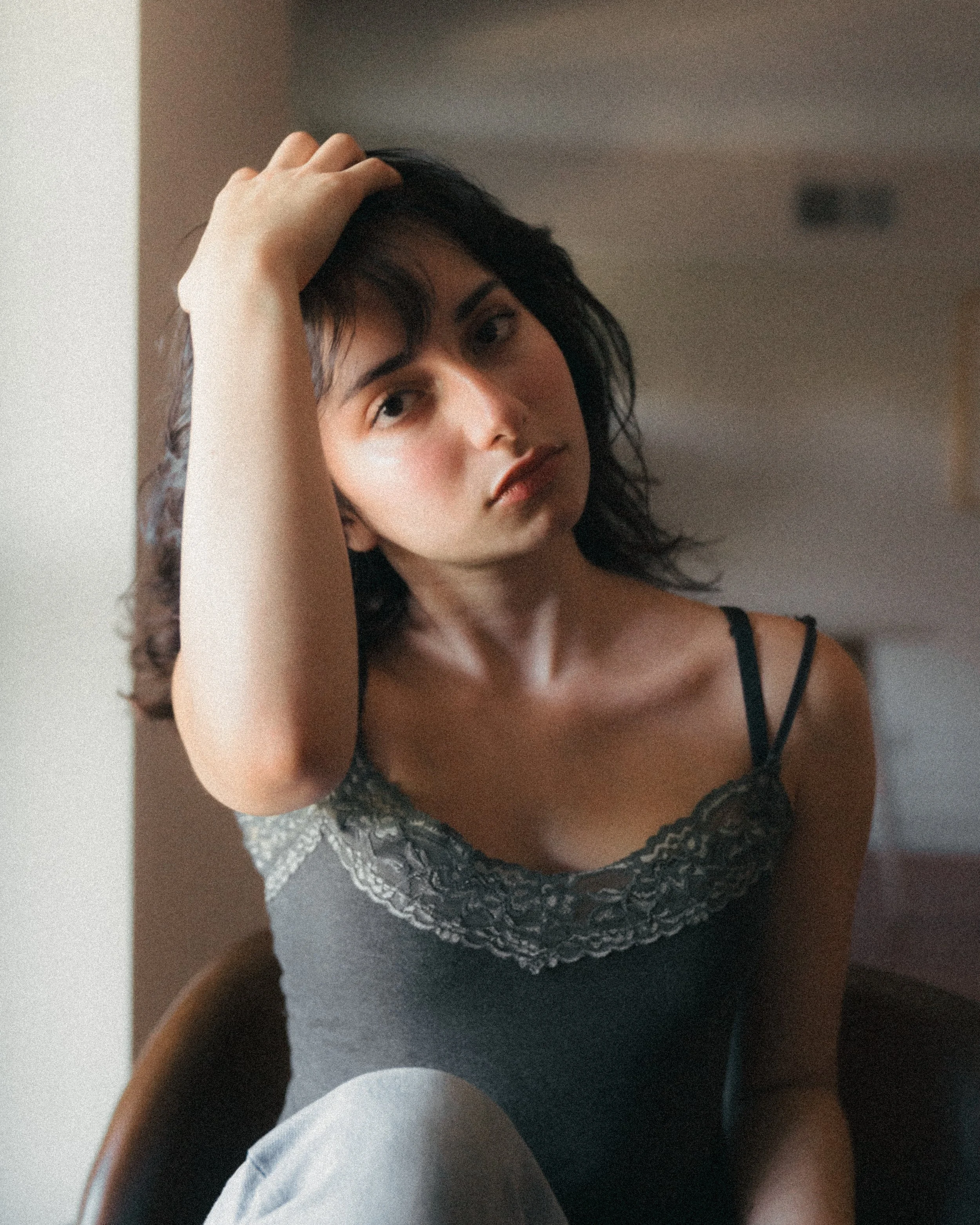 A woman with dark wavy hair, wearing a black lace-trimmed tank top, sitting on a chair, tilting her head to the side and looking into the camera with a neutral expression.