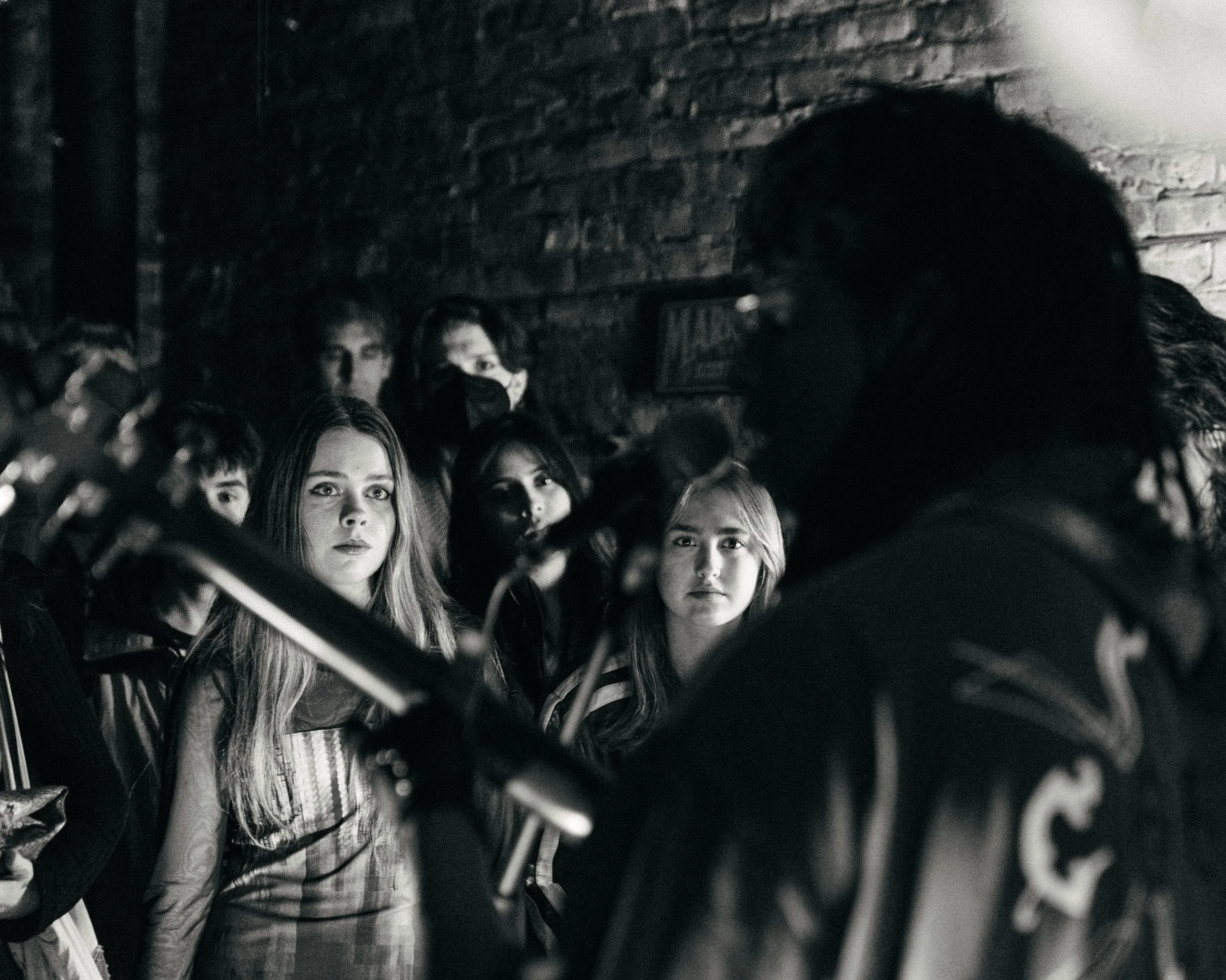 A black-and-white photo of a group of people, mostly young women, watching a performer who appears to be playing a guitar. The scene is set against a brick wall, and the lighting highlights the expressions of the audience.