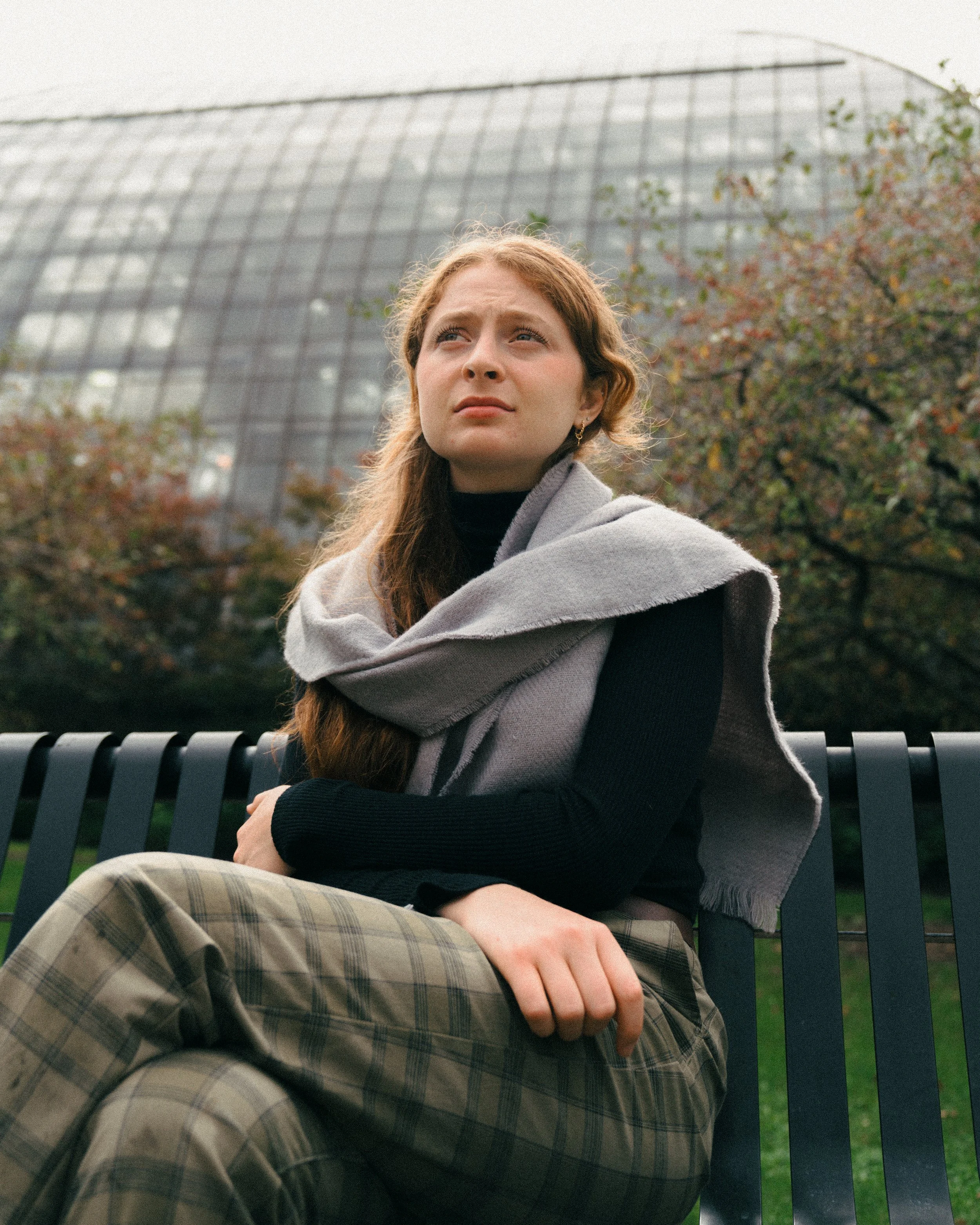 A woman with red hair sitting on a park bench, looking thoughtful, with autumn trees and modern building in the background.