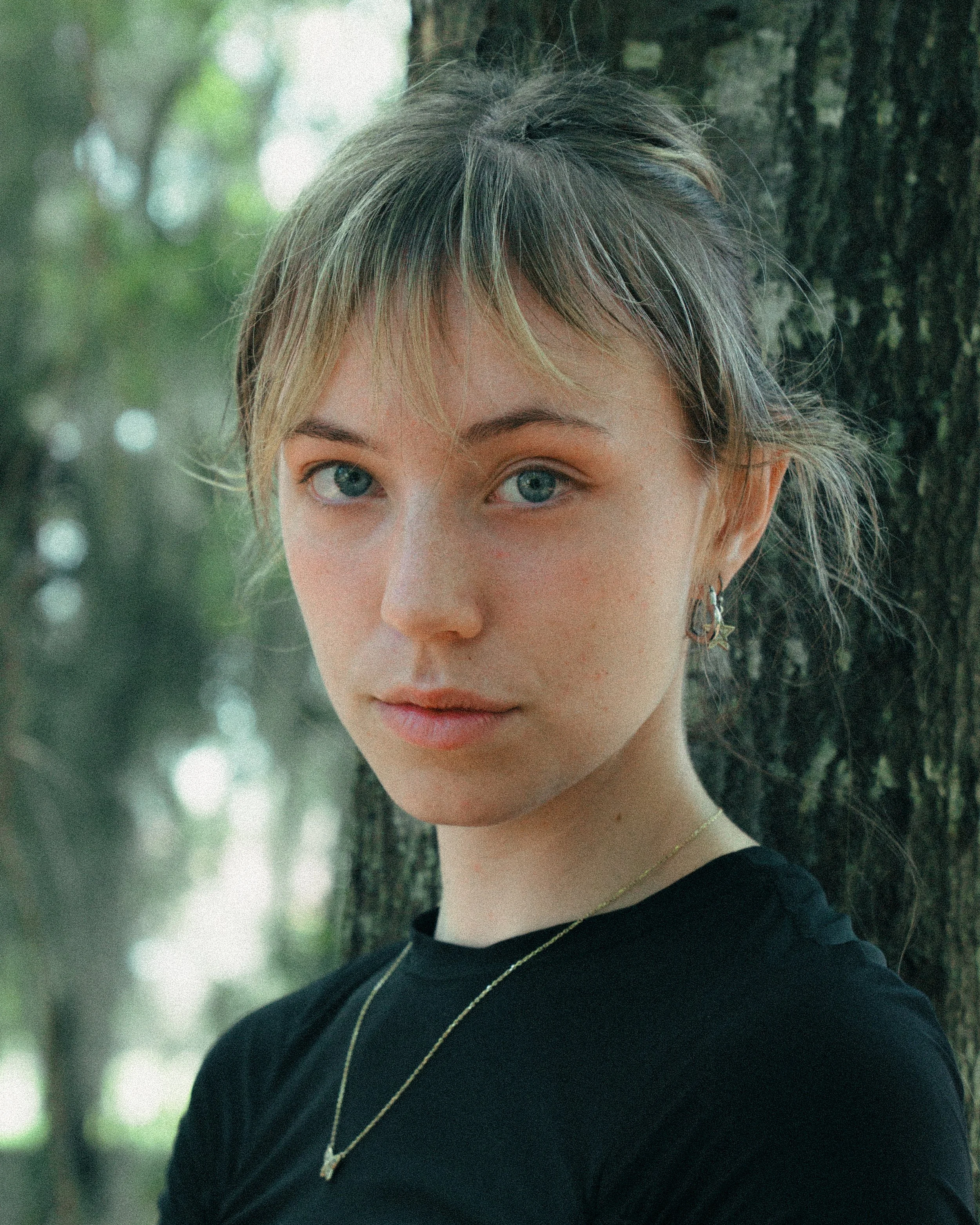 A young woman with blonde hair and blue eyes standing outdoors next to a tree, wearing a black shirt and jewelry, looking at the camera.