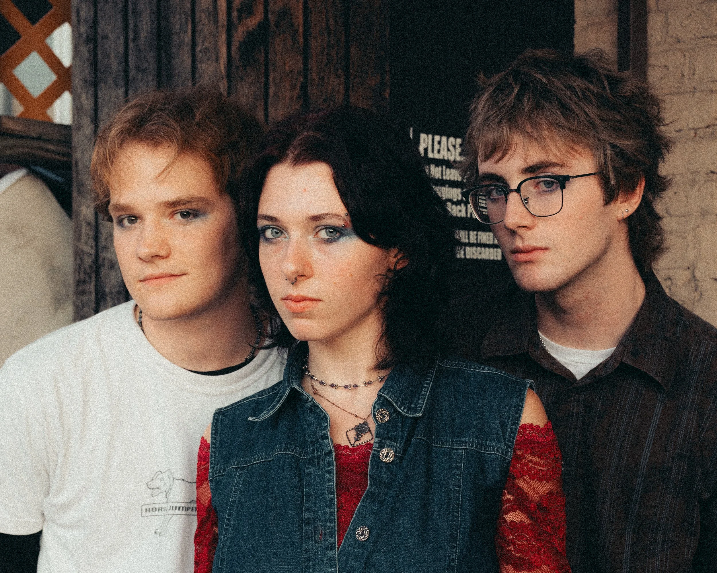 Three young individuals standing close together in a rustic setting, with a wooden background and a sign partially visible behind them.