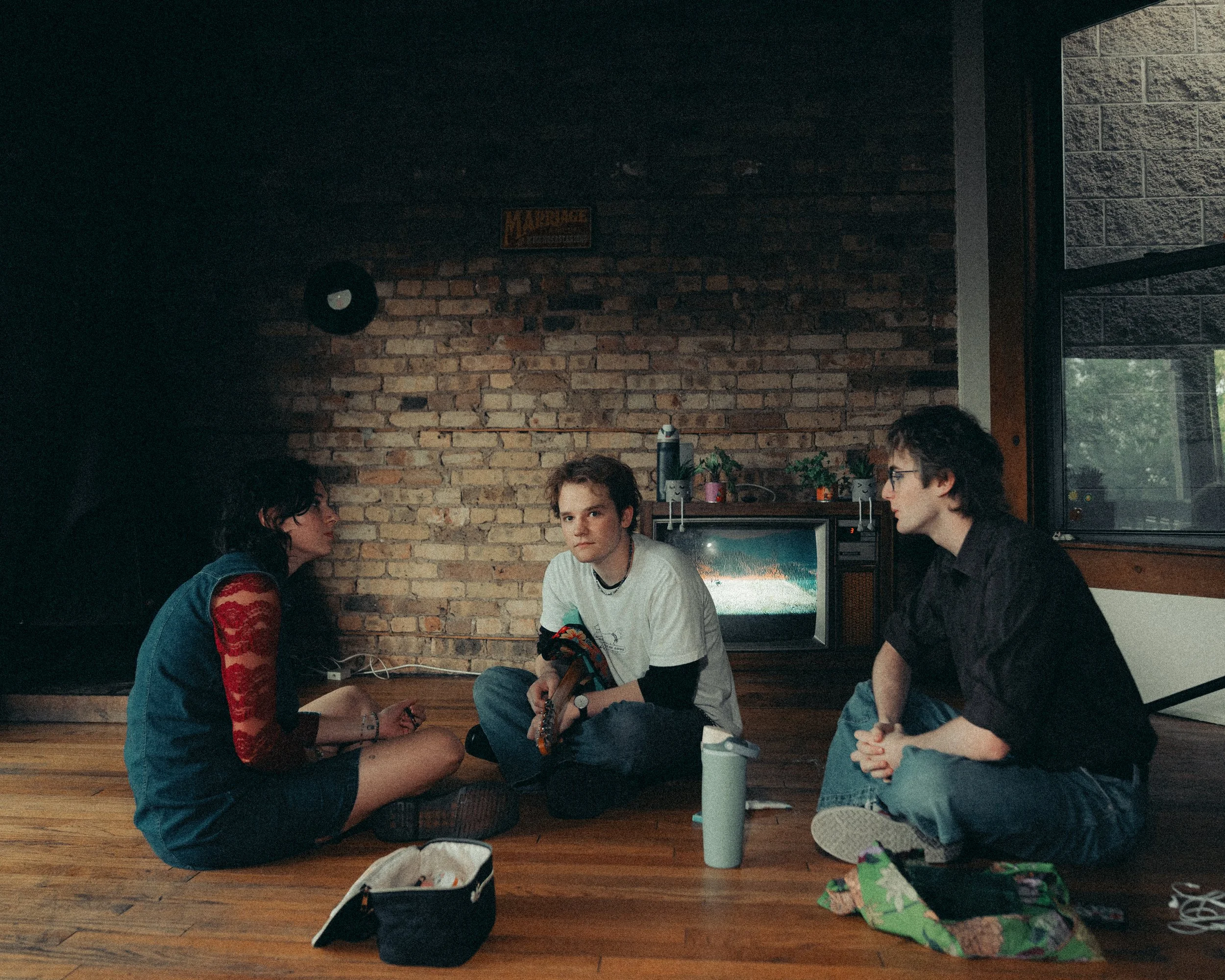 Three young people sitting on a wooden floor in a room with brick wall, two of them are facing each other, the third one is looking at the camera, with a television and potted plants behind them.