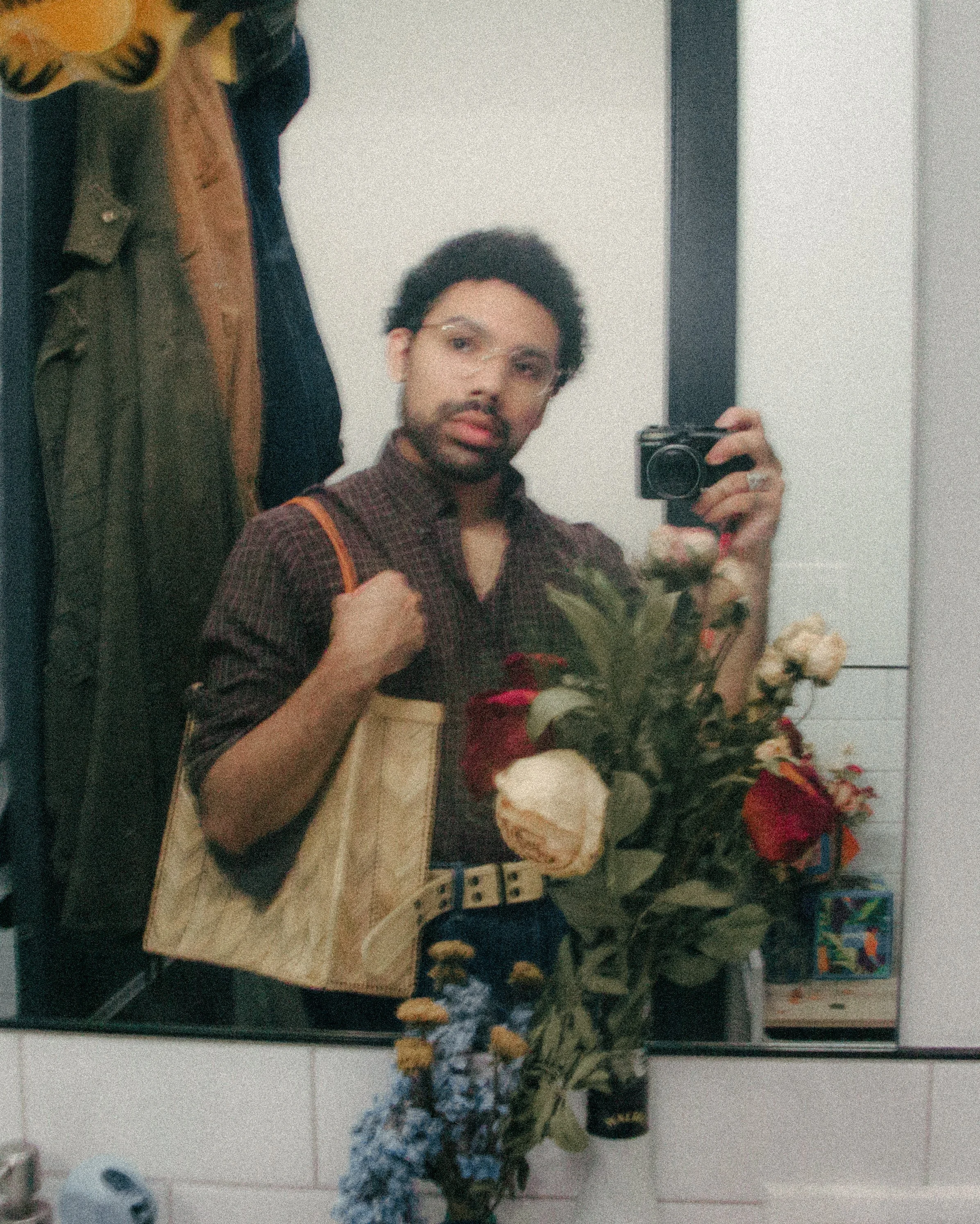 Person taking a mirror selfie in a room with hanging coats, purple shirt, glasses, and flowers in front of the mirror.