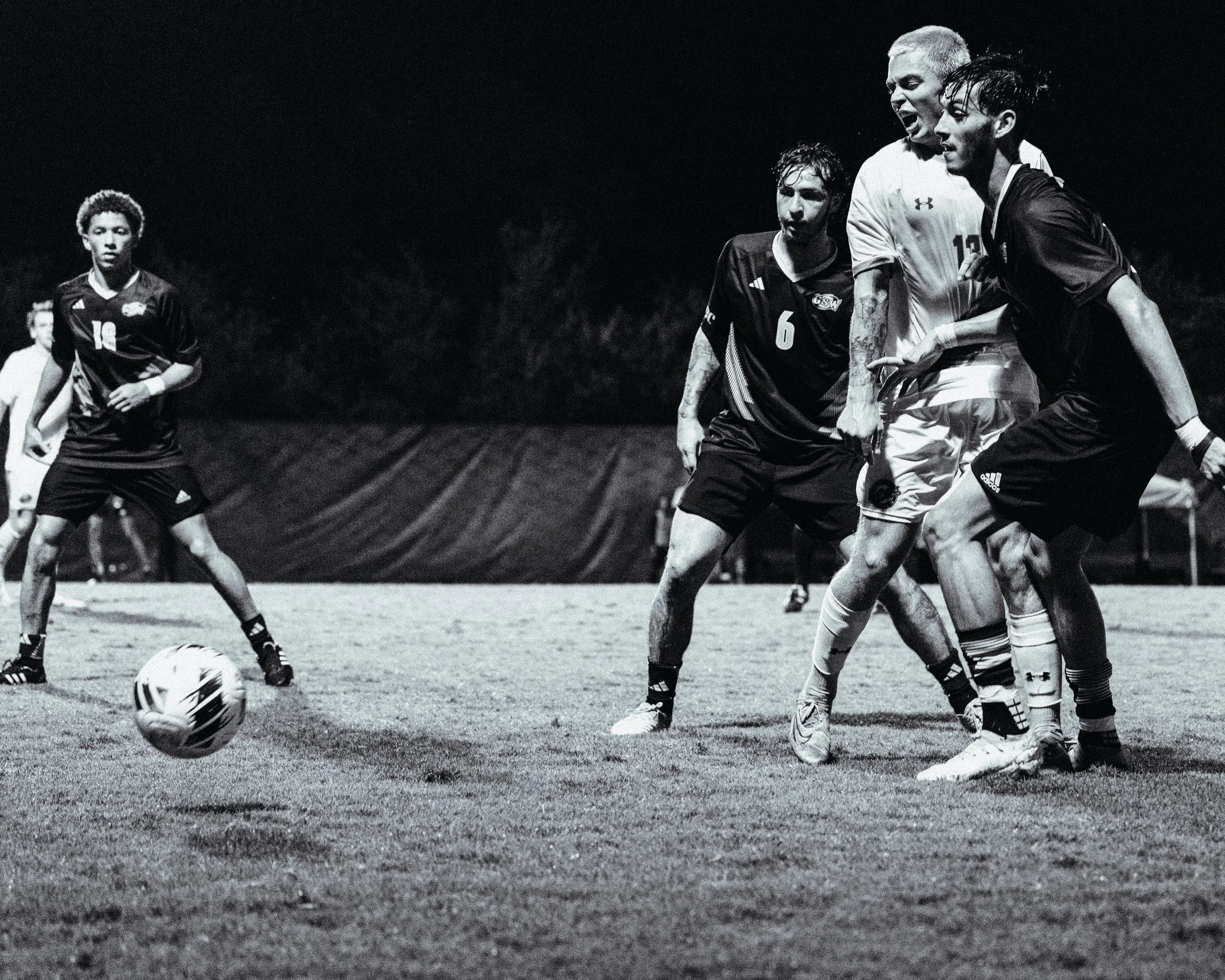 Black and white photo of a soccer game with four players, three of them appear to be fighting for the ball, on a grassy field at night.