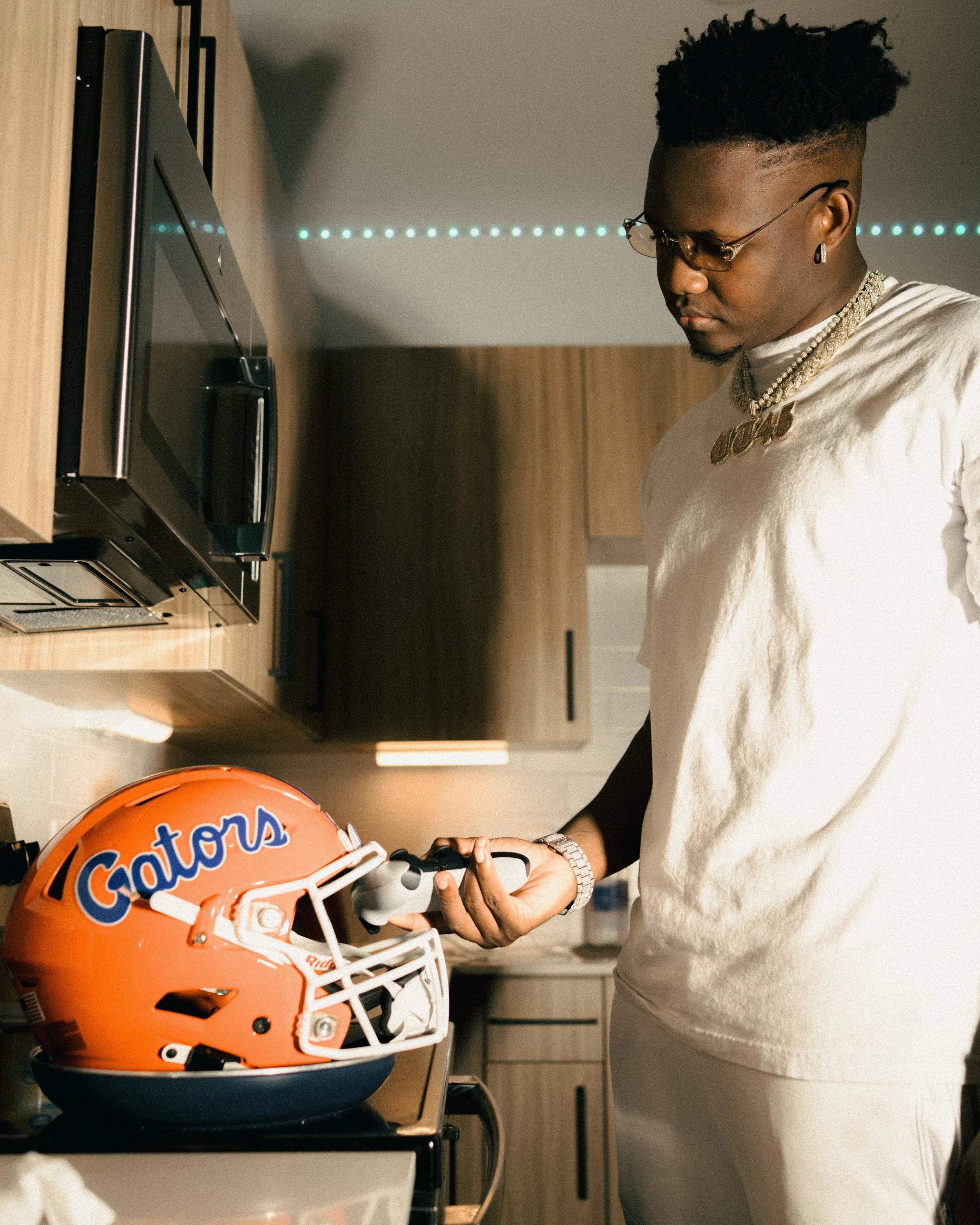 A man in white clothing and glasses holds a small electronic device next to a Gators football helmet on a kitchen counter.