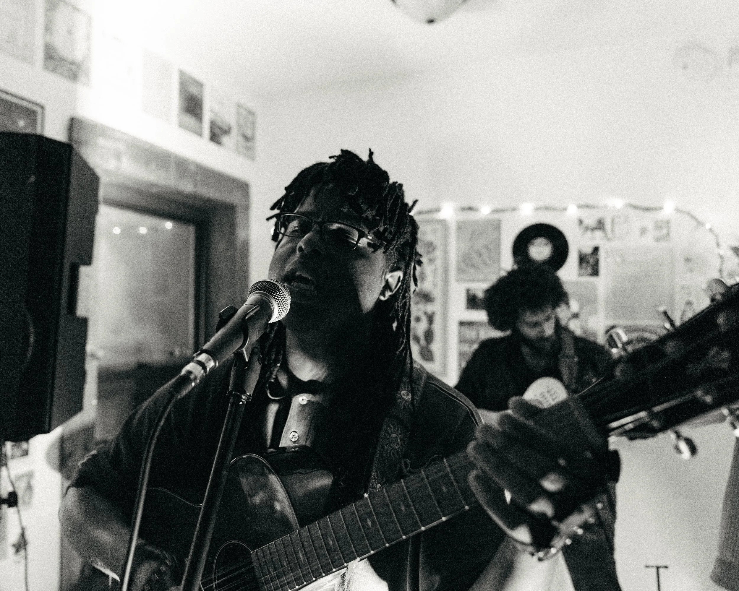 Musician with glasses singing into a microphone and playing an acoustic guitar, with another person playing guitar in the background.