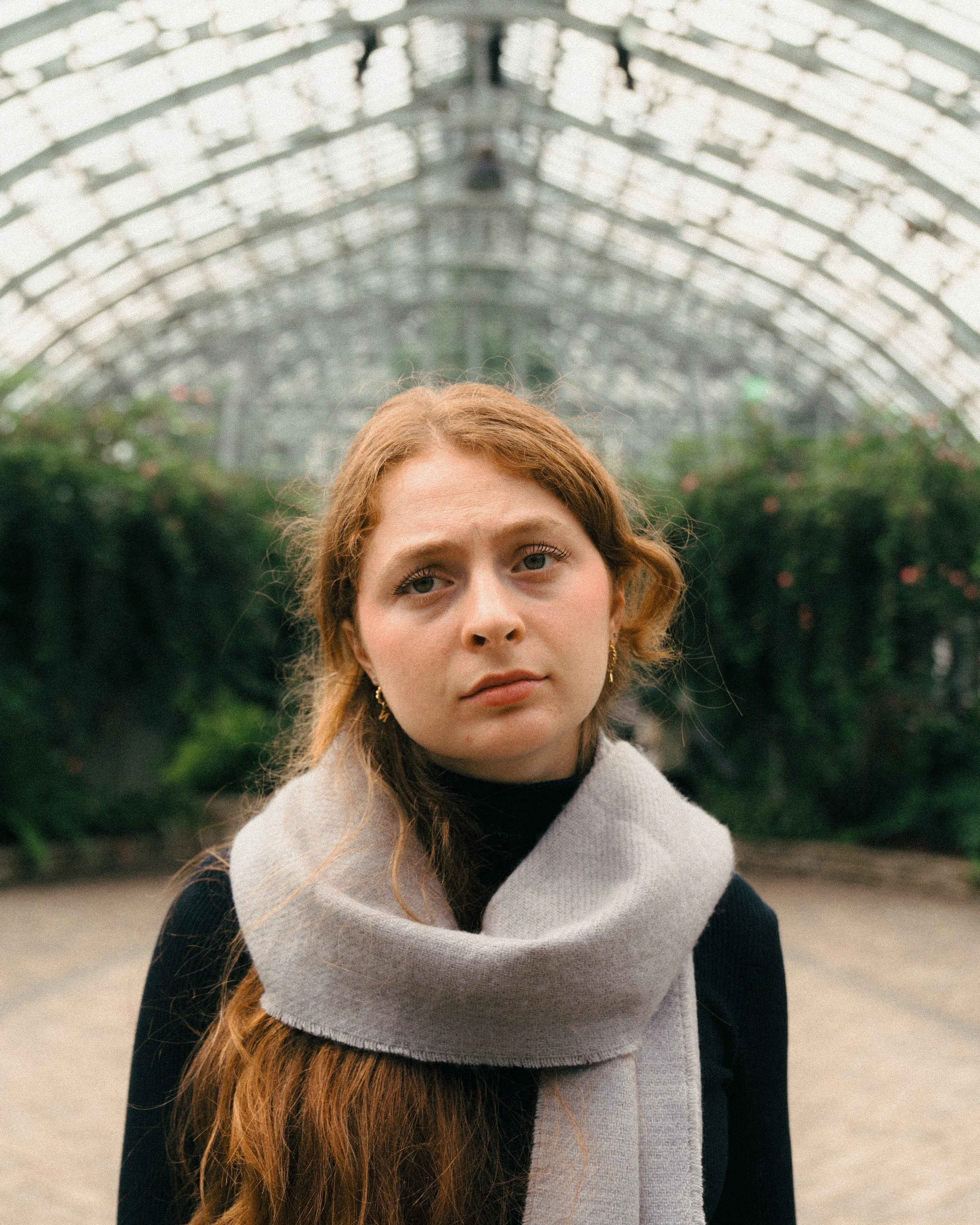 A young woman with reddish hair and fair skin stands outdoors in front of a glass and metal arched greenhouse. She is wearing a black turtleneck and a beige scarf, and has gold earrings. She looks slightly to the camera with a neutral expression.