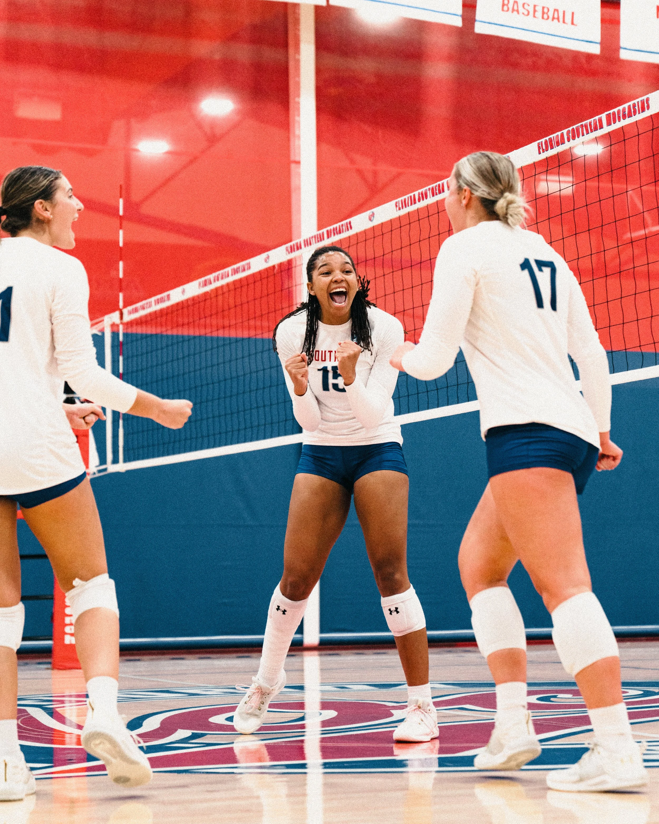 Female volleyball players celebrating on indoor court, with one player in the center showing excitement, fists clenched, and teammates around her also happy. Volleyball net visible in the background.
