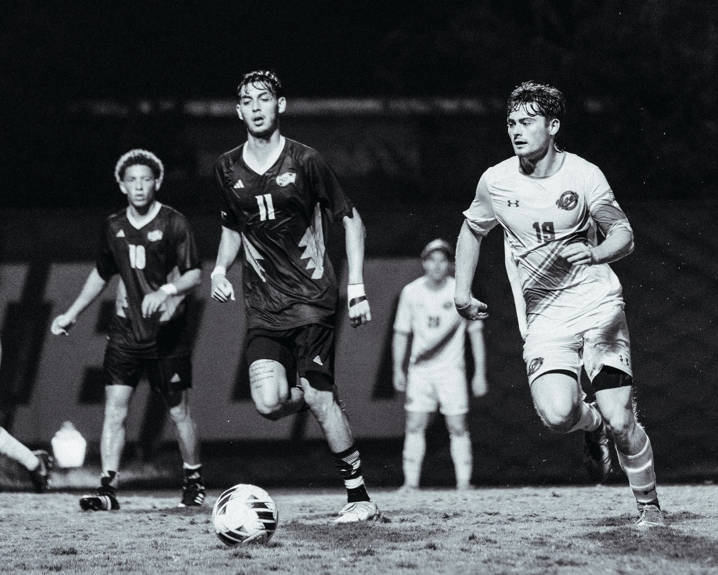 Black and white photo of a soccer match with four players on the field, two of whom are running towards the camera, with a soccer ball on the ground in front of them. Two additional players are in the background, standing and watching the game.