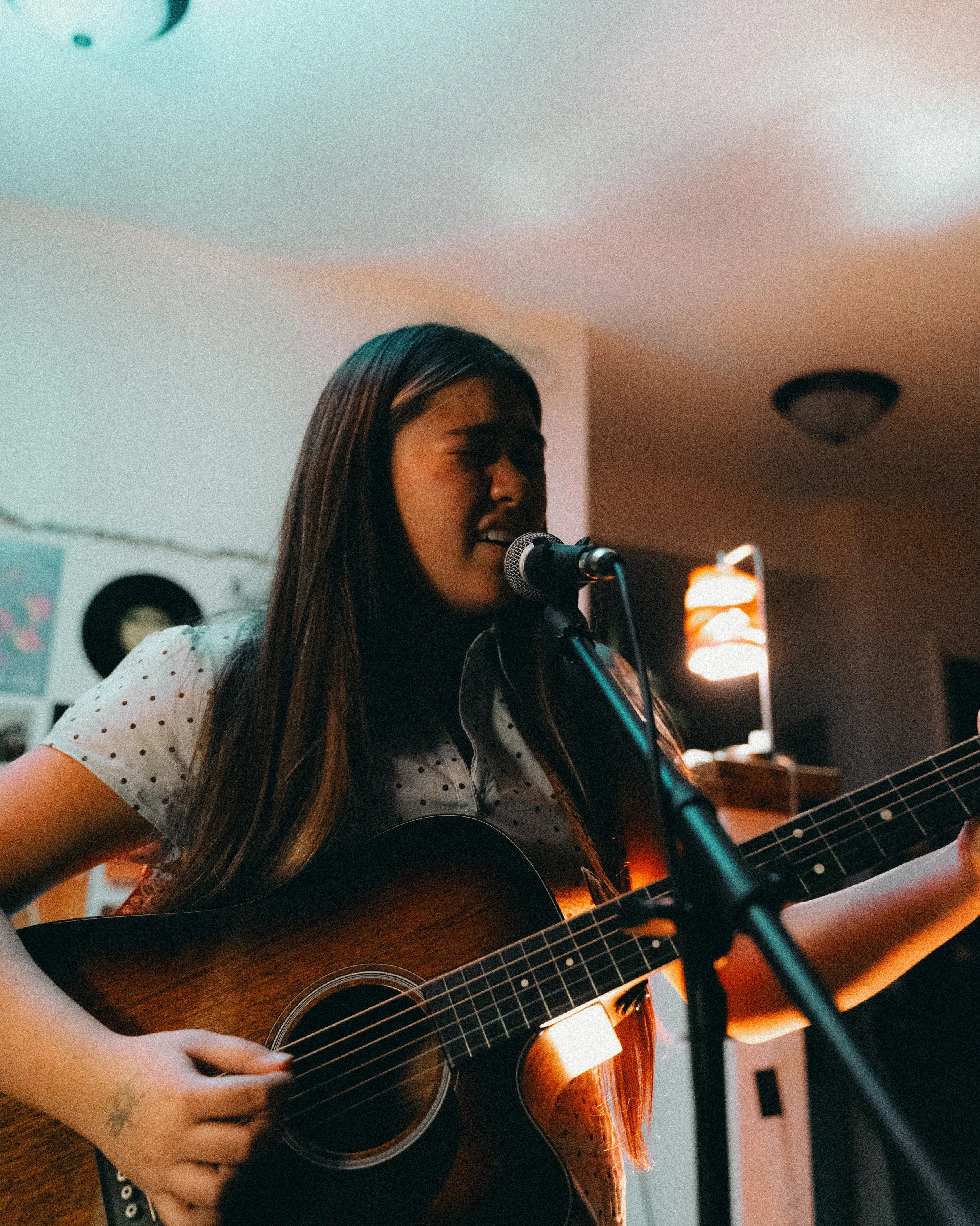 Young woman singing and playing acoustic guitar in a room with warm lighting.