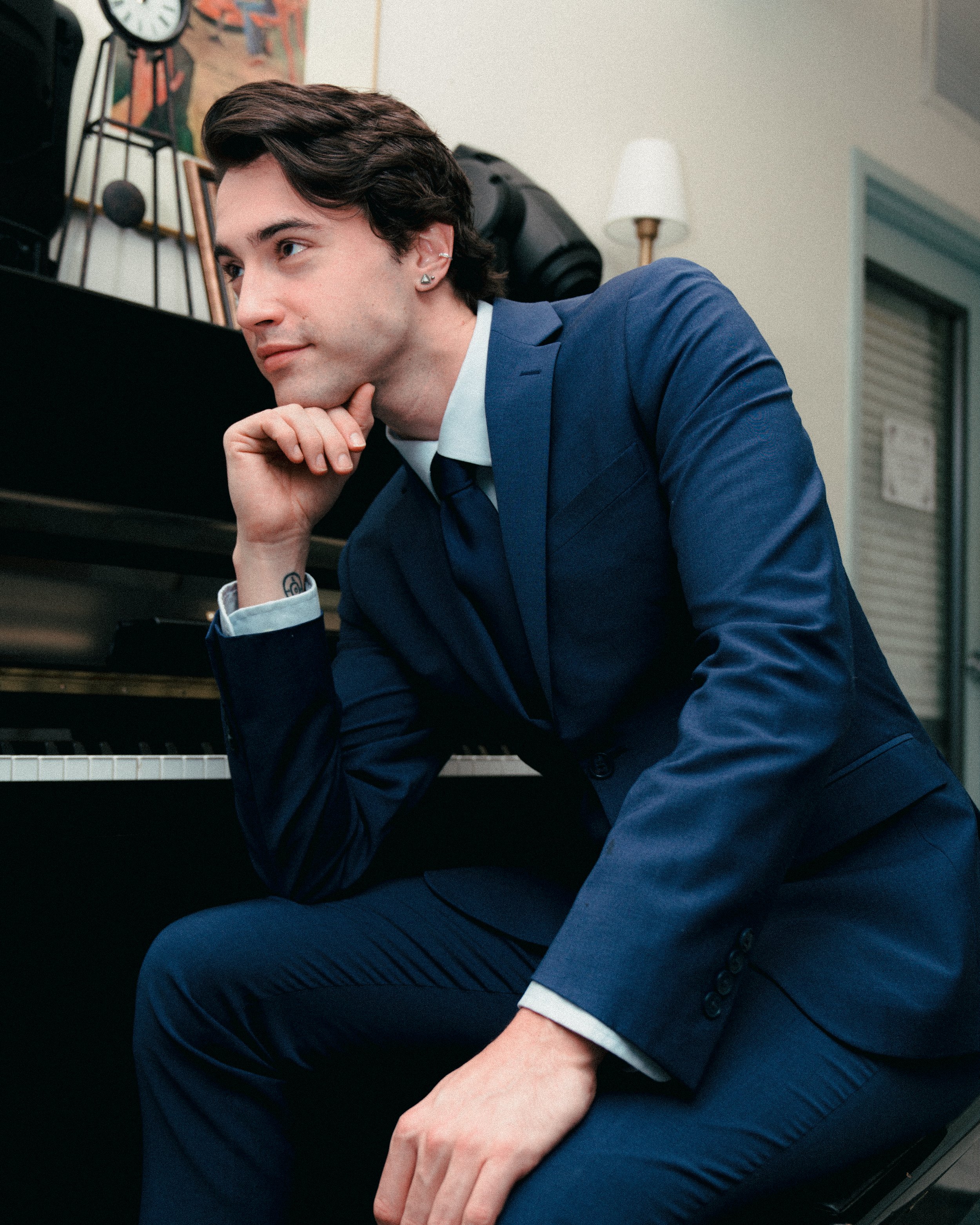 A young man in a blue suit sitting on a piano bench, resting his chin on his hand, inside a room with a painting, a clock, and a white lamp in the background.
