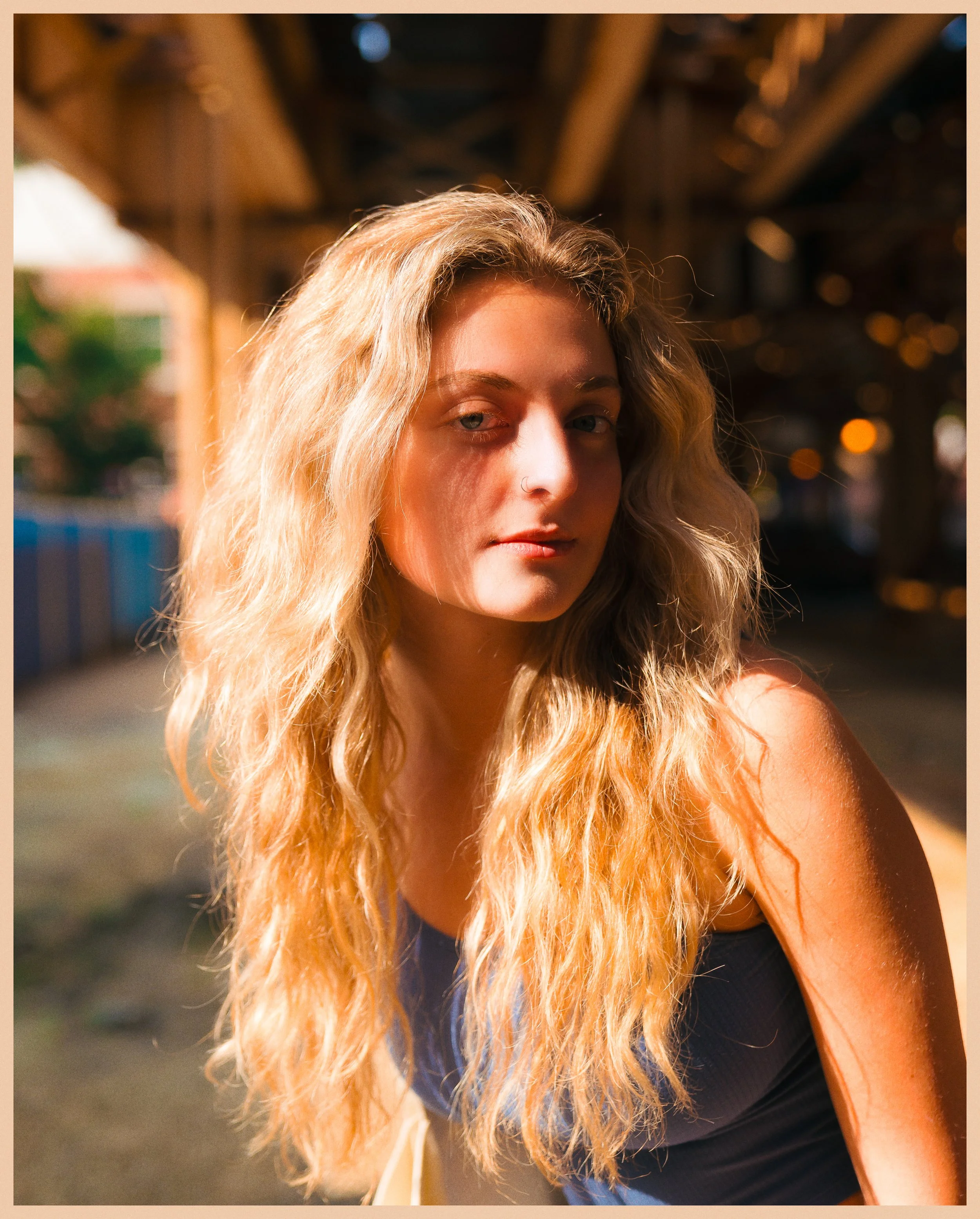 A young woman with long, wavy blonde hair and a nose piercing, standing outdoors during golden hour, with a blurred background of a structure and string lights.