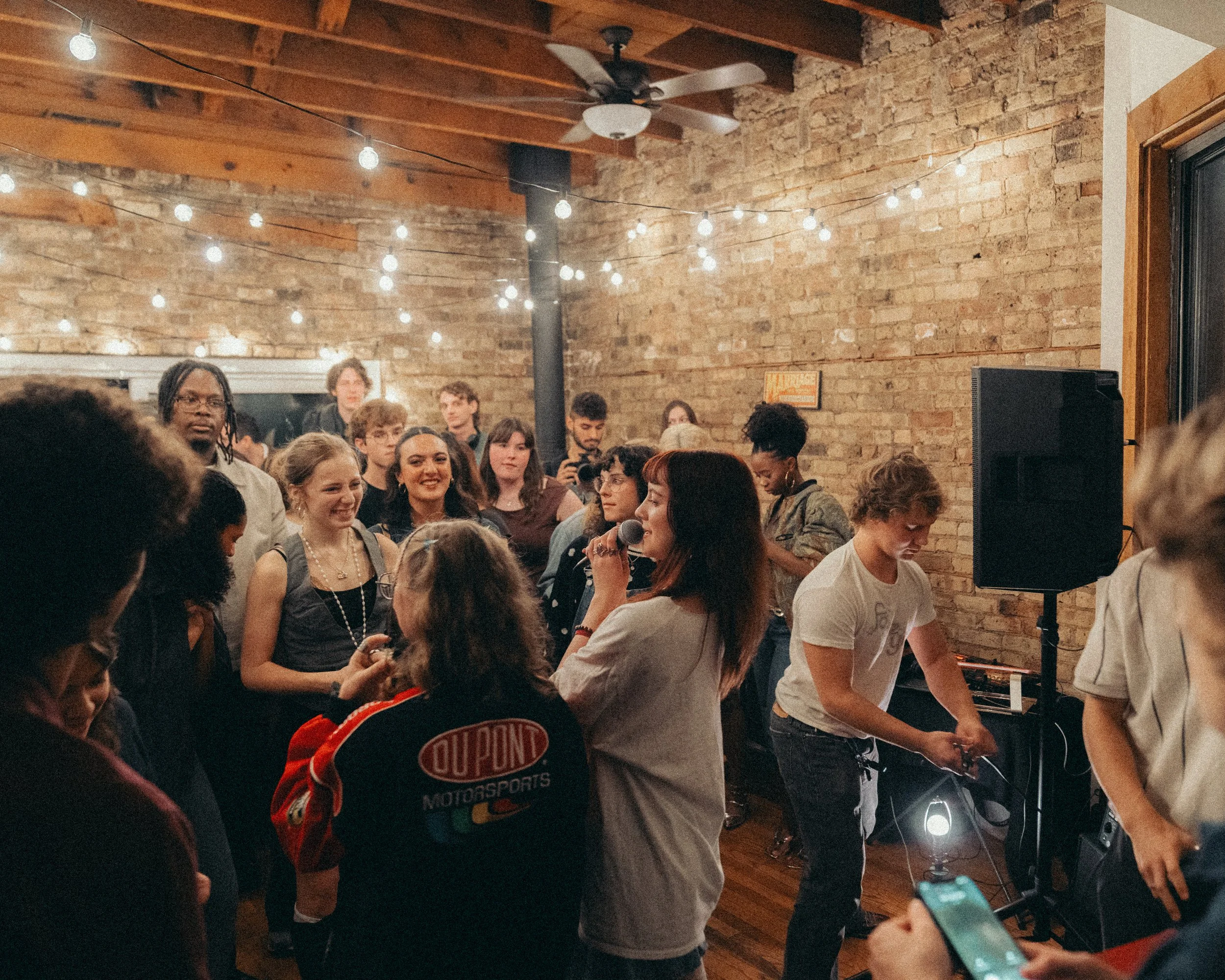 A group of people gathered in a room with exposed brick walls and string lights hanging from the ceiling. A woman is speaking into a microphone, and another woman appears to be adjusting sound equipment. The crowd is smiling and enjoying the event.