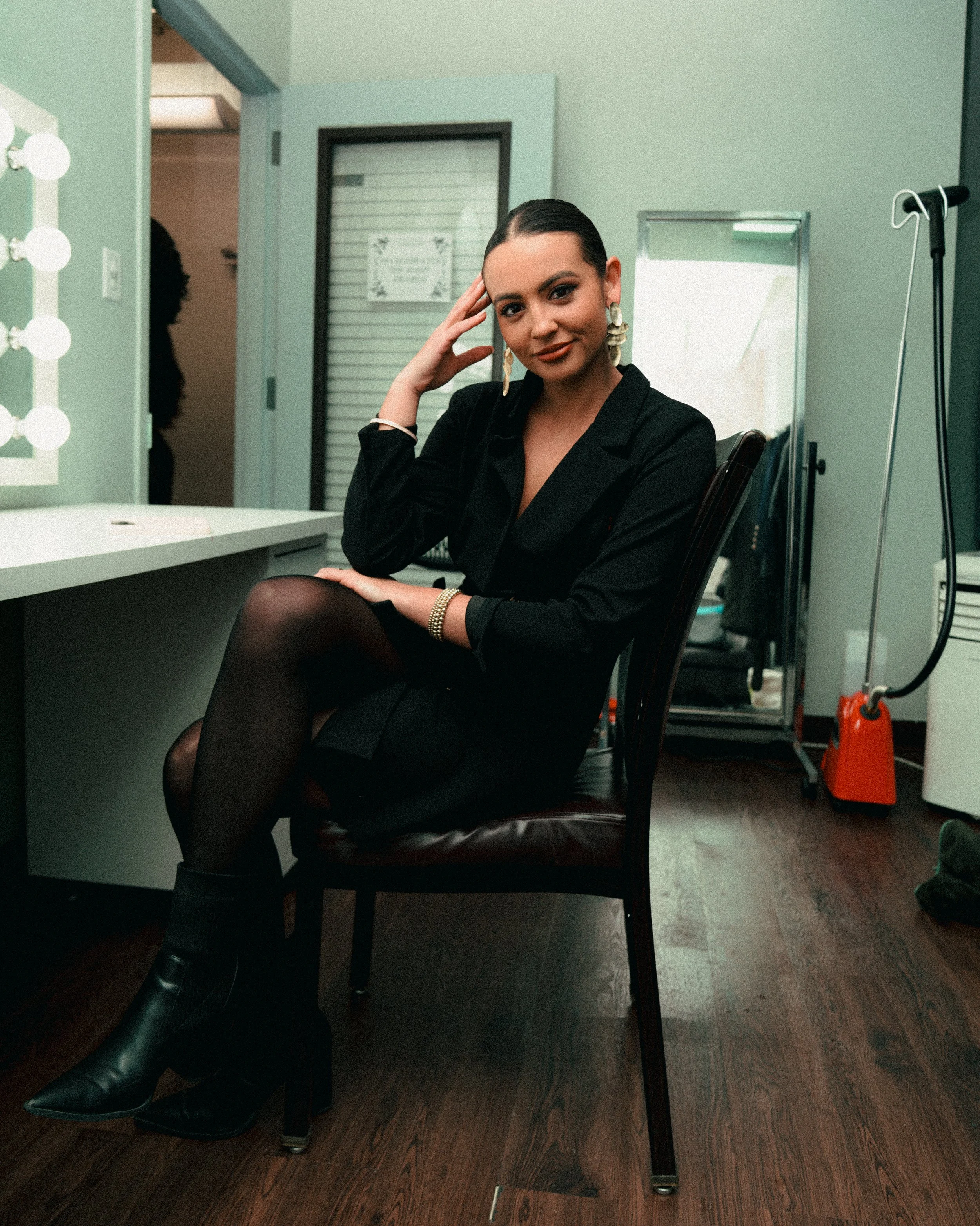 A woman sitting on a chair at a dressing room vanity, dressed in black, wearing black tights and boots, with earrings and bracelet, posing with her hand on her temple and a slight smile.
