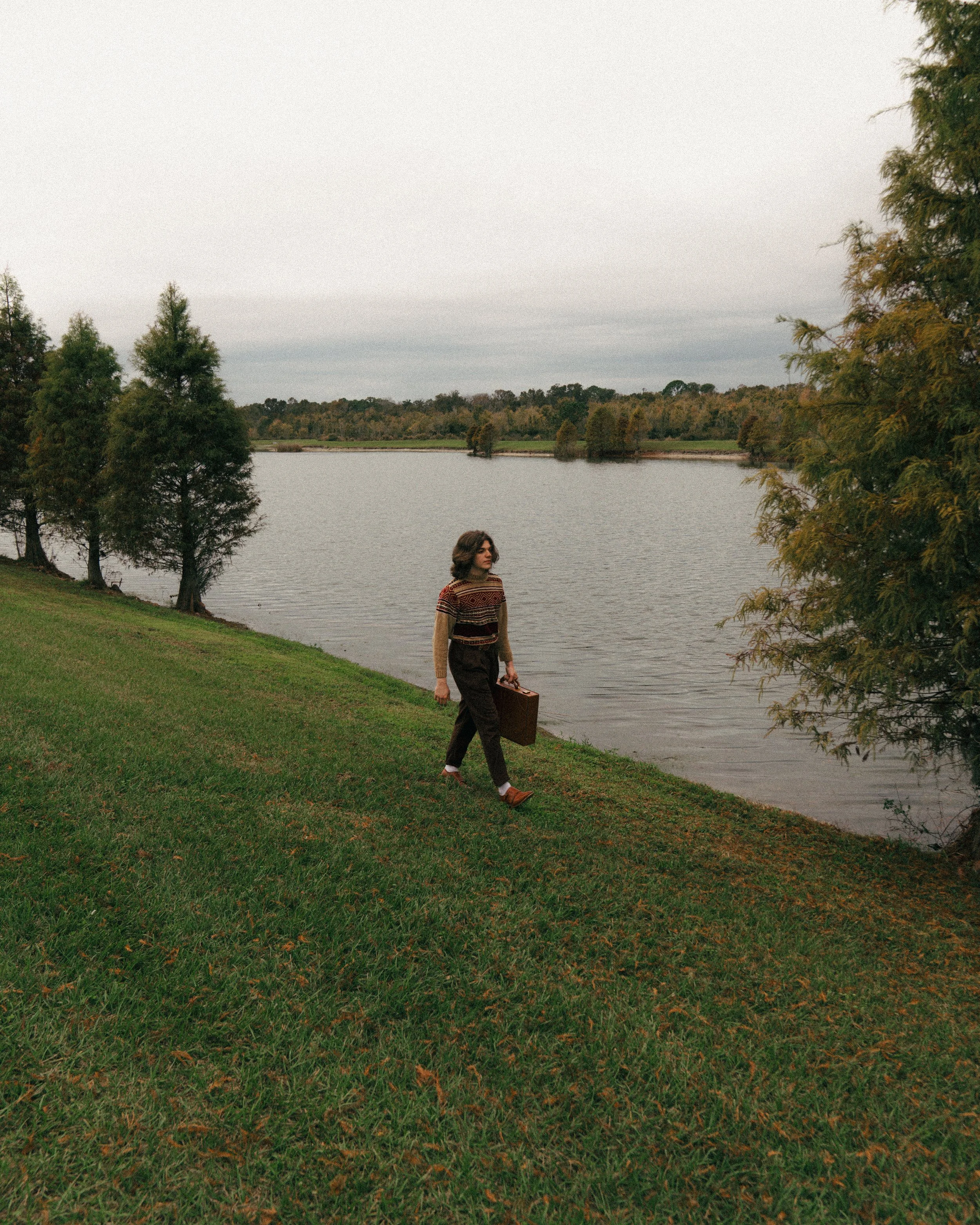 A woman walks along the bank of a lake carrying a vintage suitcase during overcast weather in a park with green grass and trees.