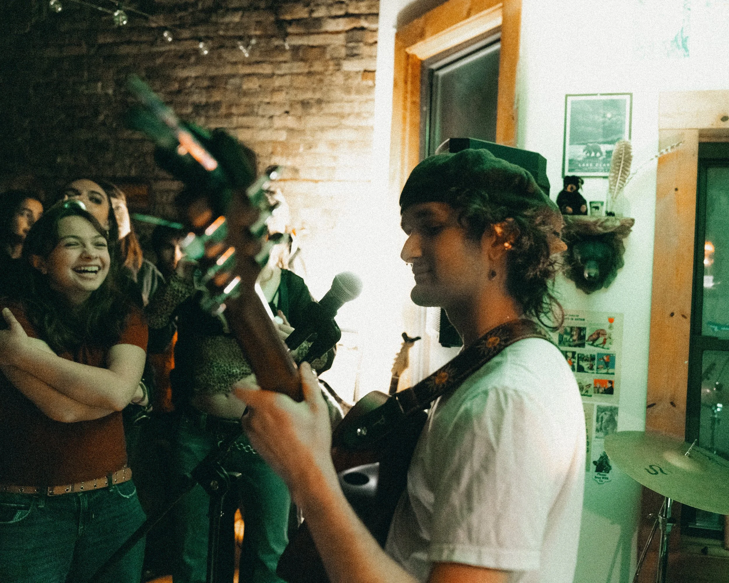 A woman with curly hair and a green beret playing an acoustic guitar, performing in front of an audience of young women smiling and enjoying the music inside a cozy venue with brick and wooden walls, decorated with pictures and artwork.