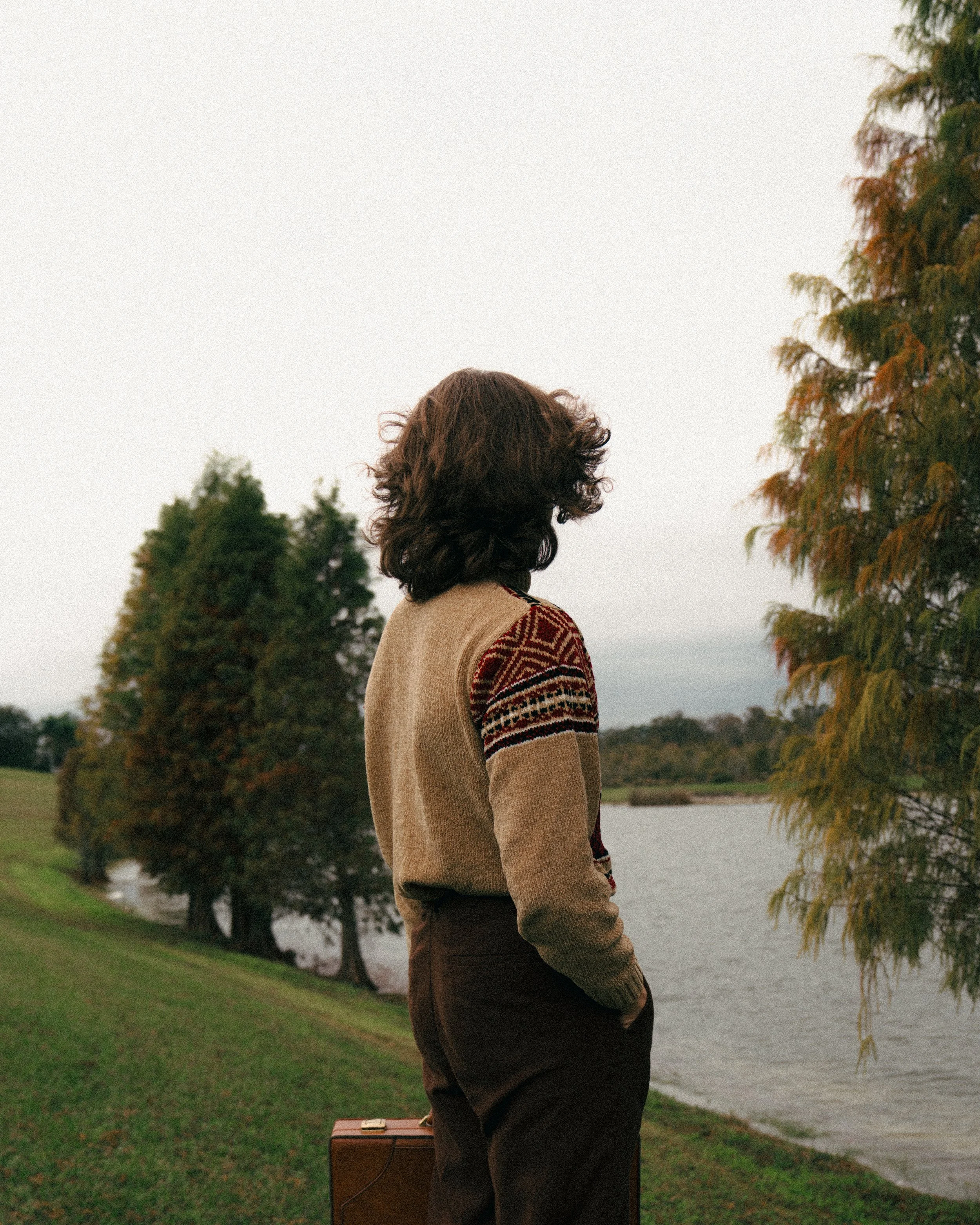 A person with shoulder-length brown curly hair standing outdoors near a body of water, with trees in the background and wearing a patterned sweater and dark pants.