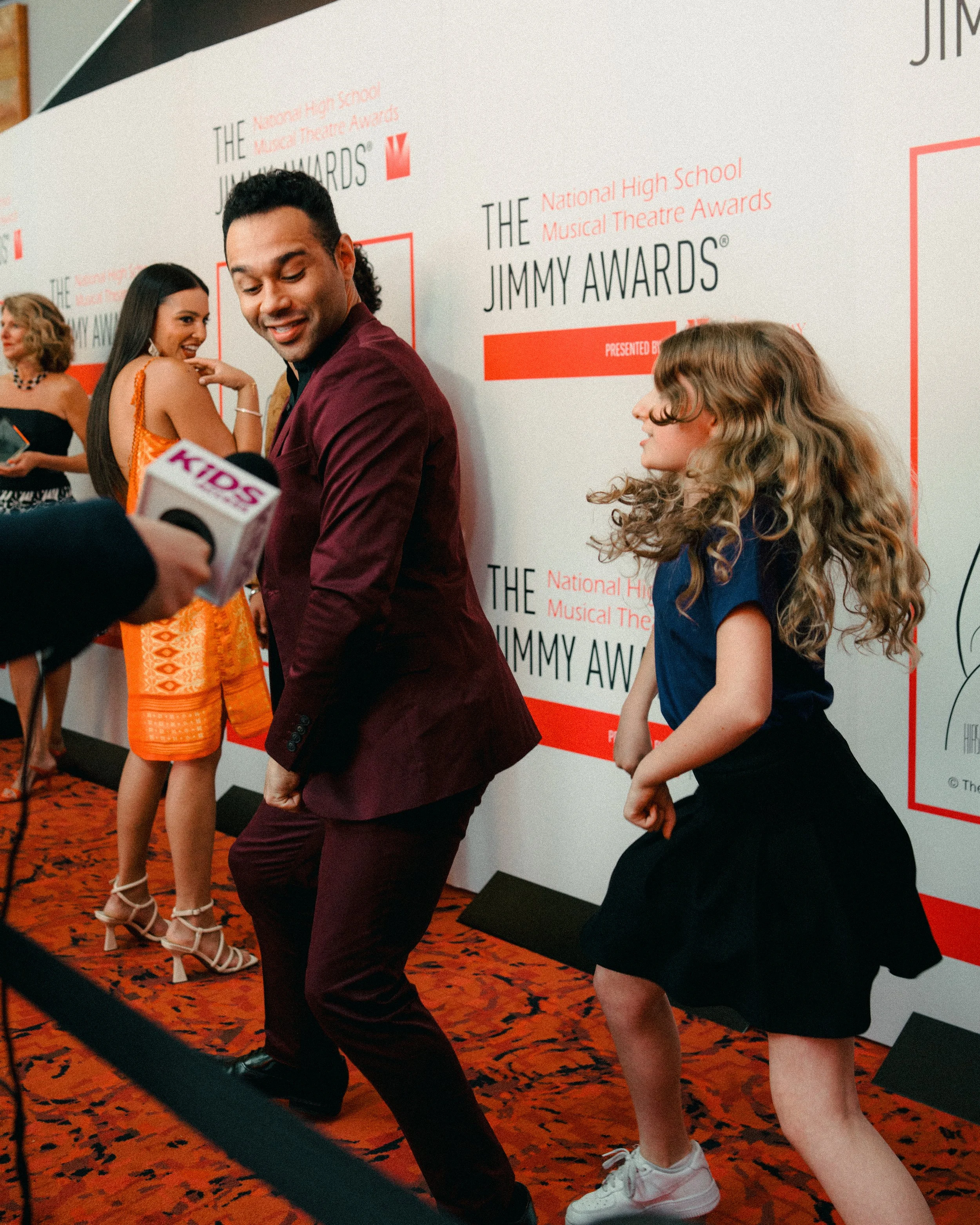 A group of people at the Jimmy Awards, with a man and young girl in the foreground. The man is smiling and standing in front of a backdrop that reads 'The Jimmy Awards,' and a woman in a colorful dress is in the background.