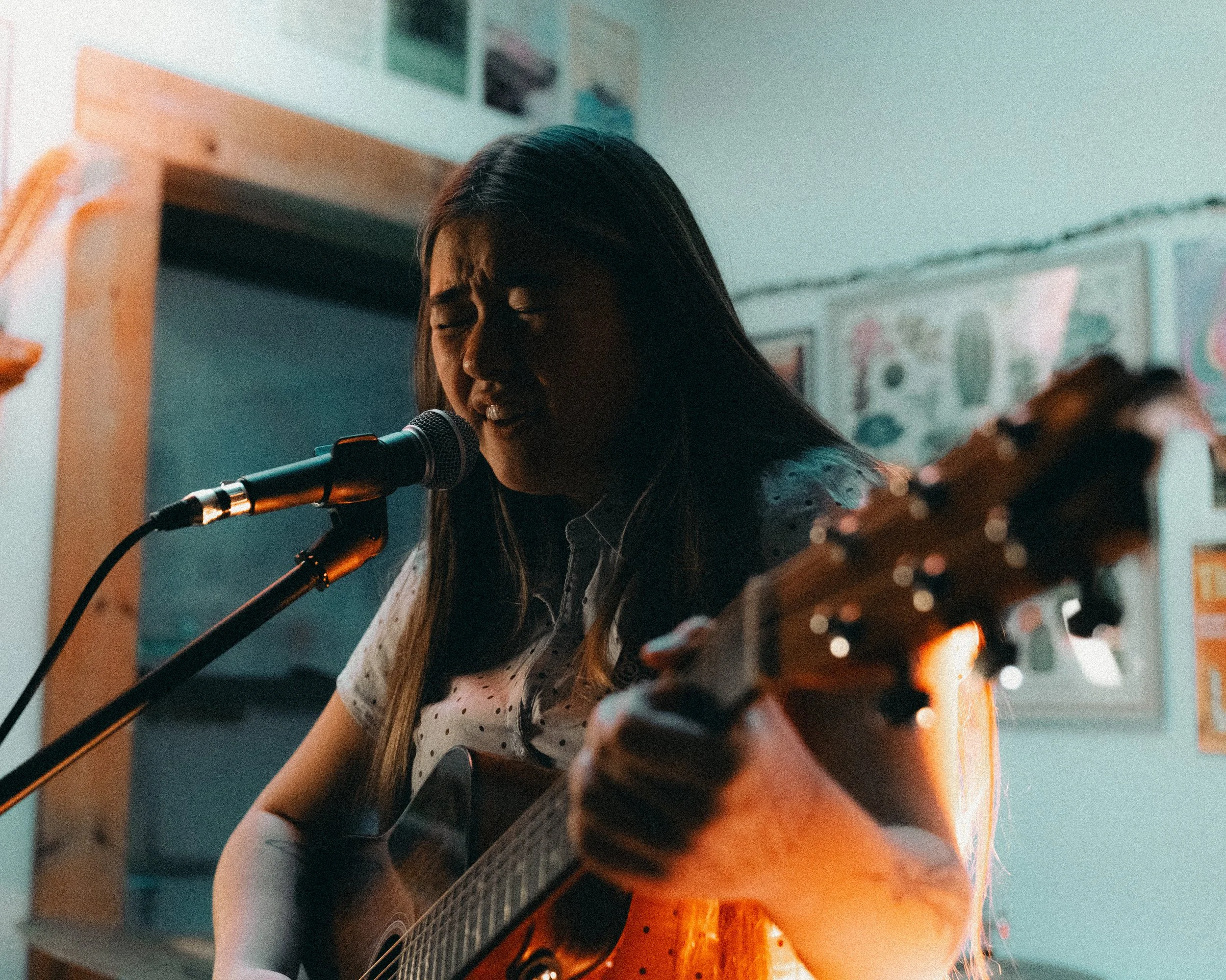 Young woman singing into a microphone while playing an acoustic guitar in a room decorated with posters and artwork.