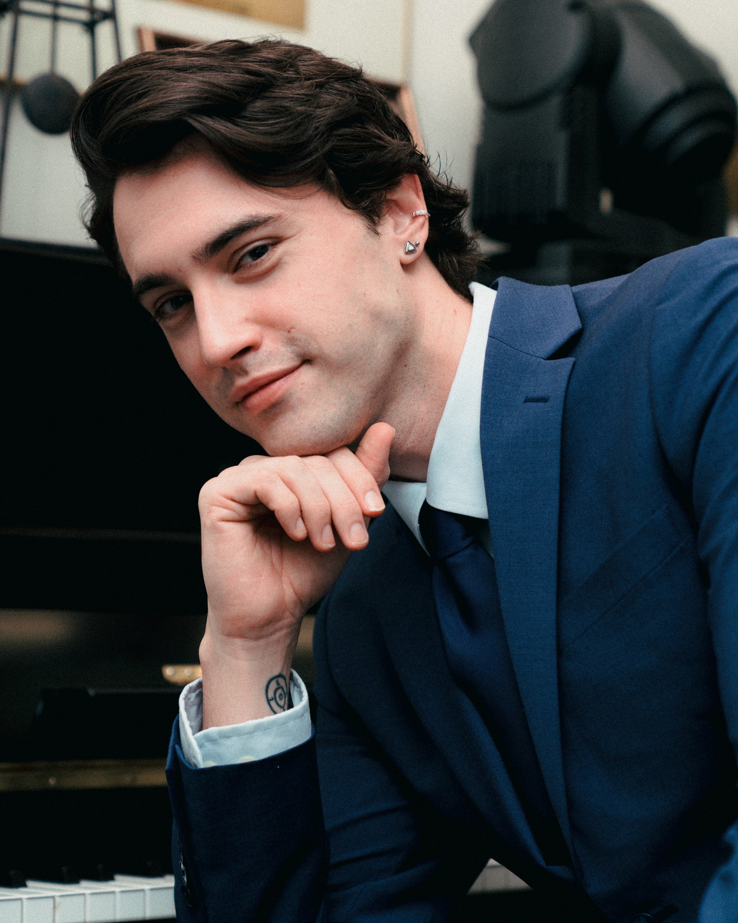 A young man with dark, wavy hair, wearing a blue suit and white shirt, resting his chin on his hand and looking at the camera with a slight smile, sitting in front of a piano.
