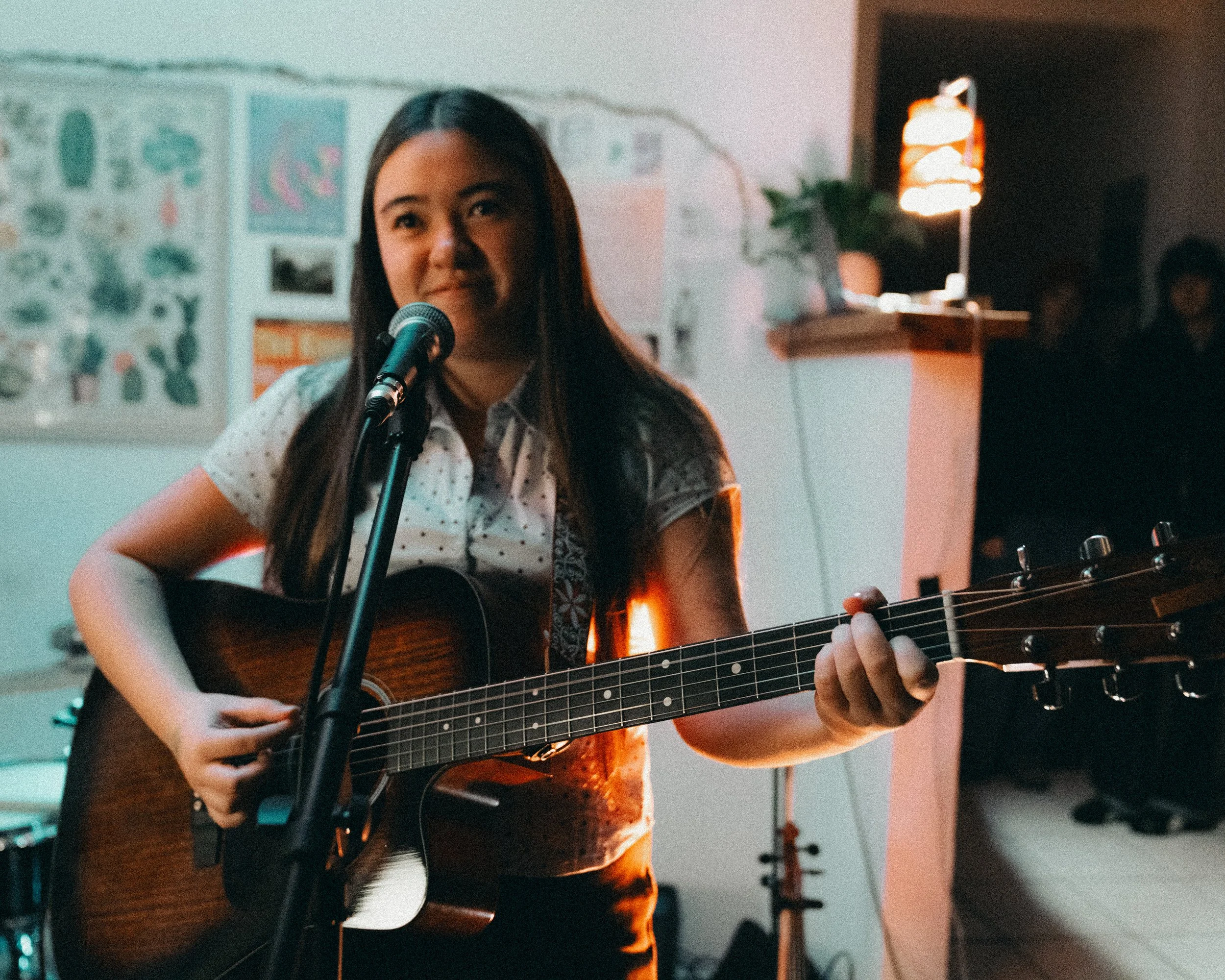 Young woman performing music with an acoustic guitar and singing into a microphone in a dimly lit room.